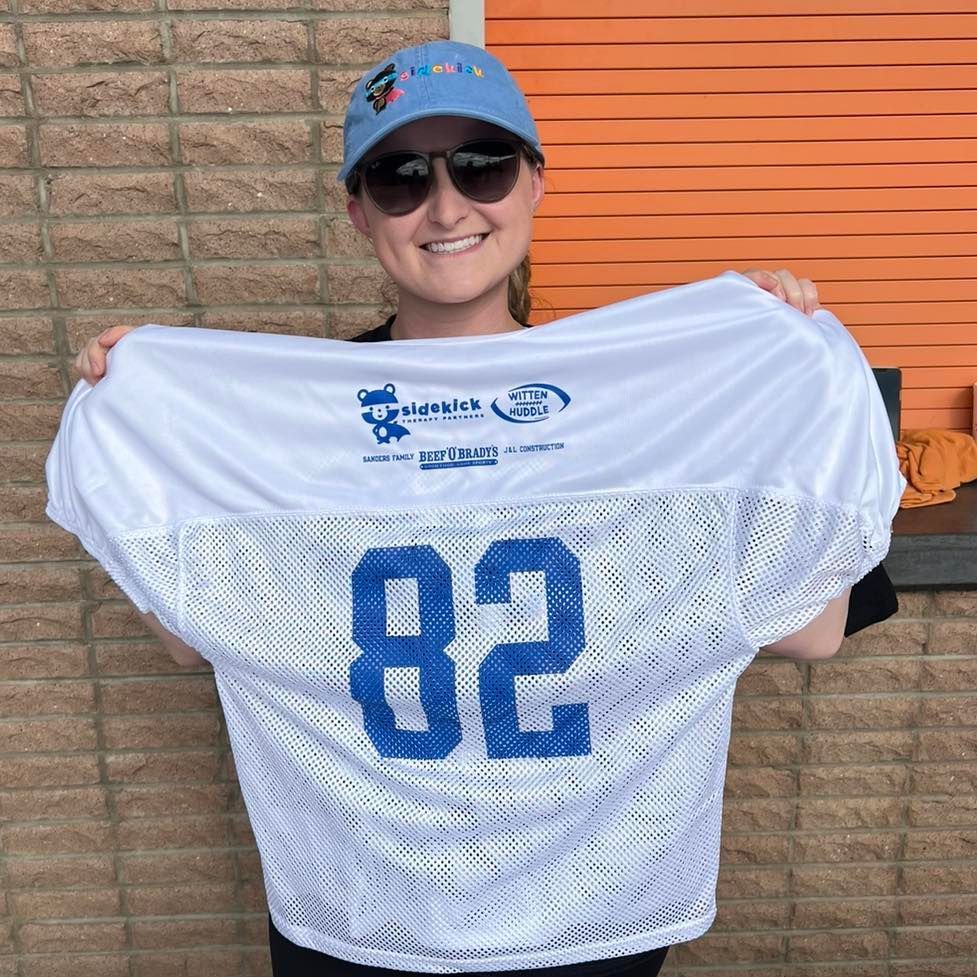 Woman holding up white football jersey with the number 82 in front of orange wall.