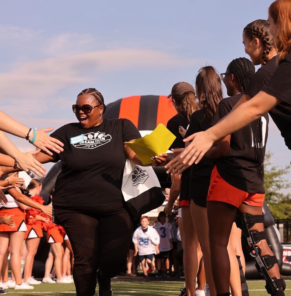 Woman with papers high-fiving students on a sports field, near a team entrance arch.