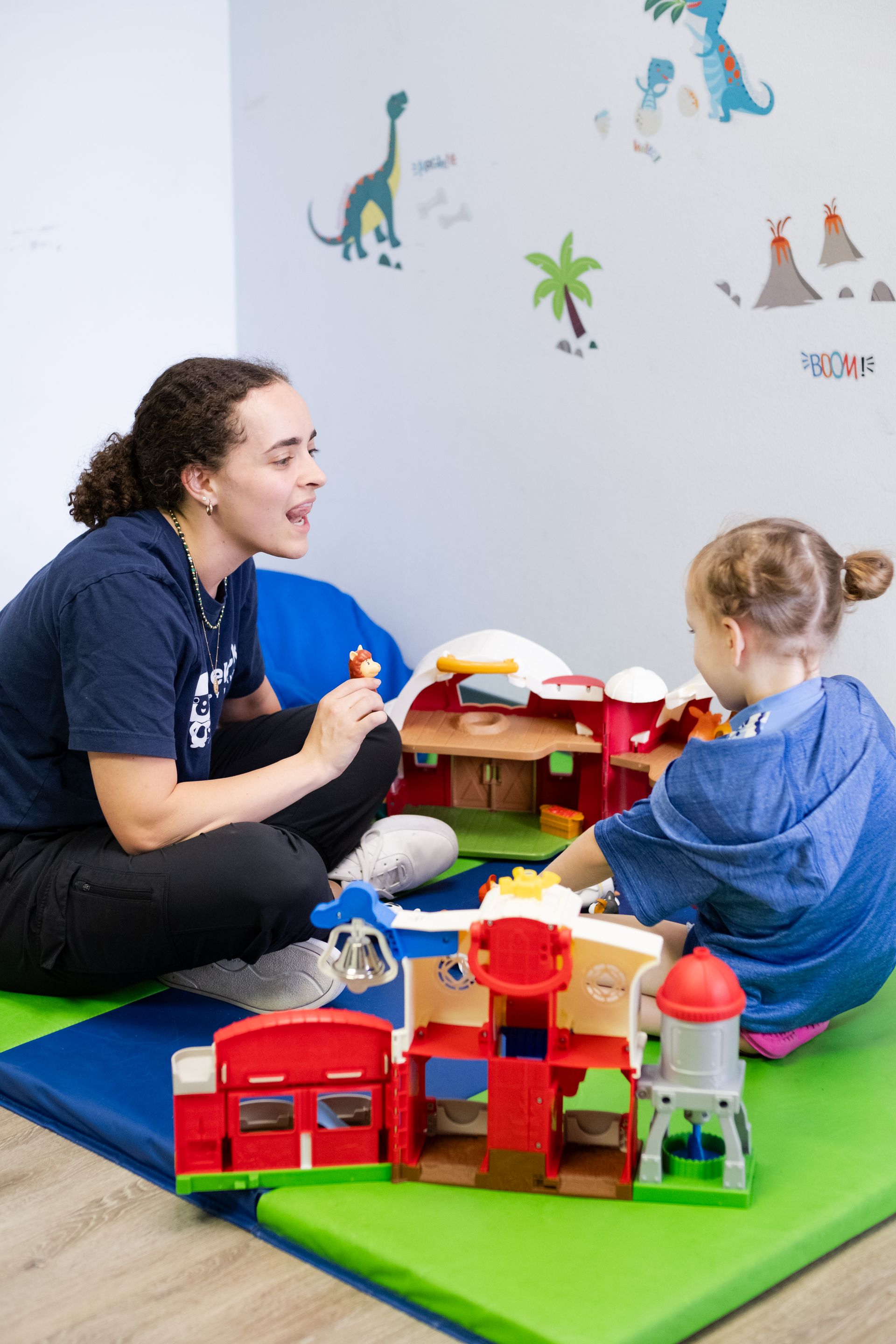 Woman and child playing with toys in a room. Dinosaurs and palm tree on the wall.
