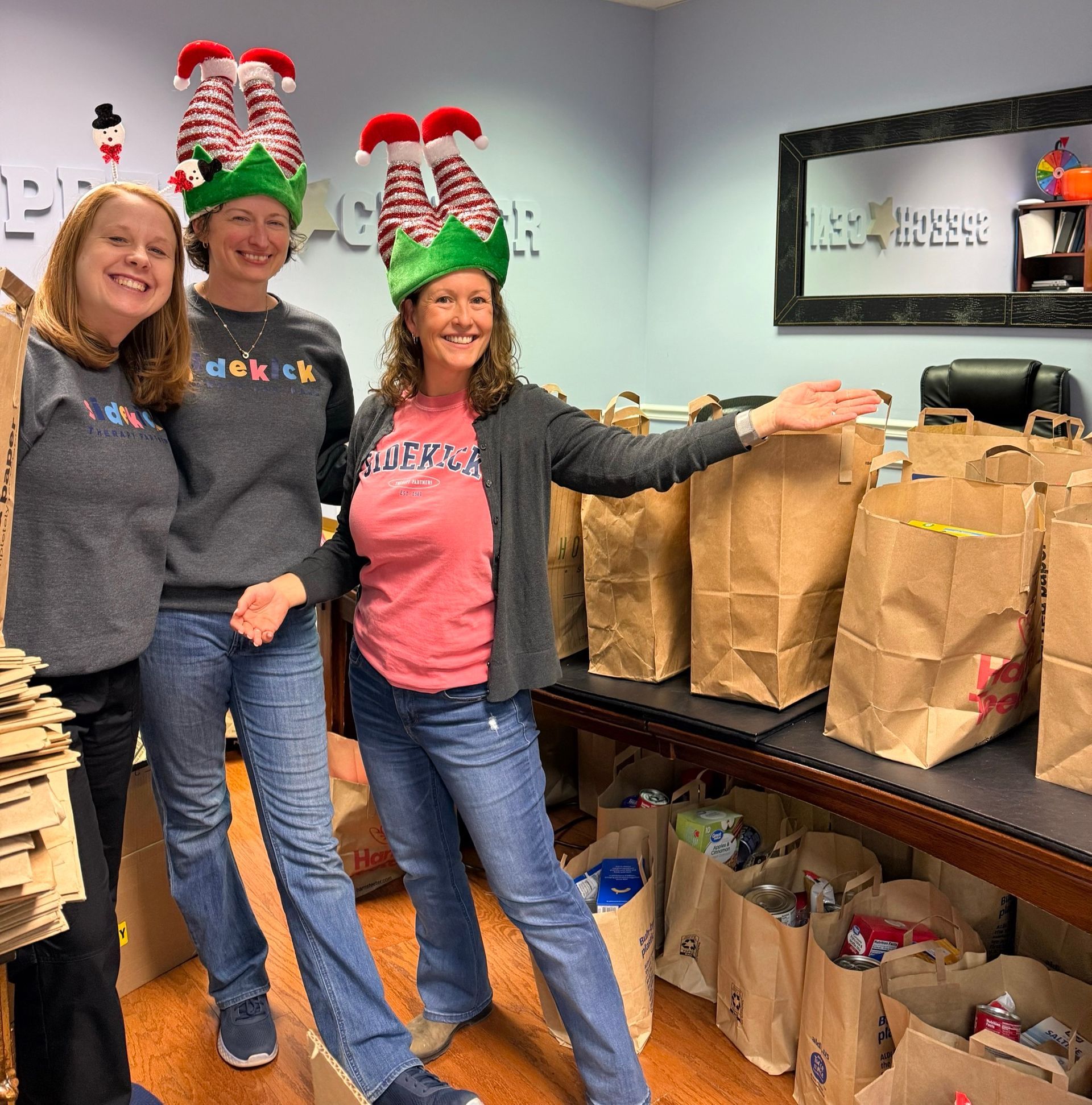 Three women wearing elf hats pose with bags of food in a room with a sign, smiling, assisting with a food drive.