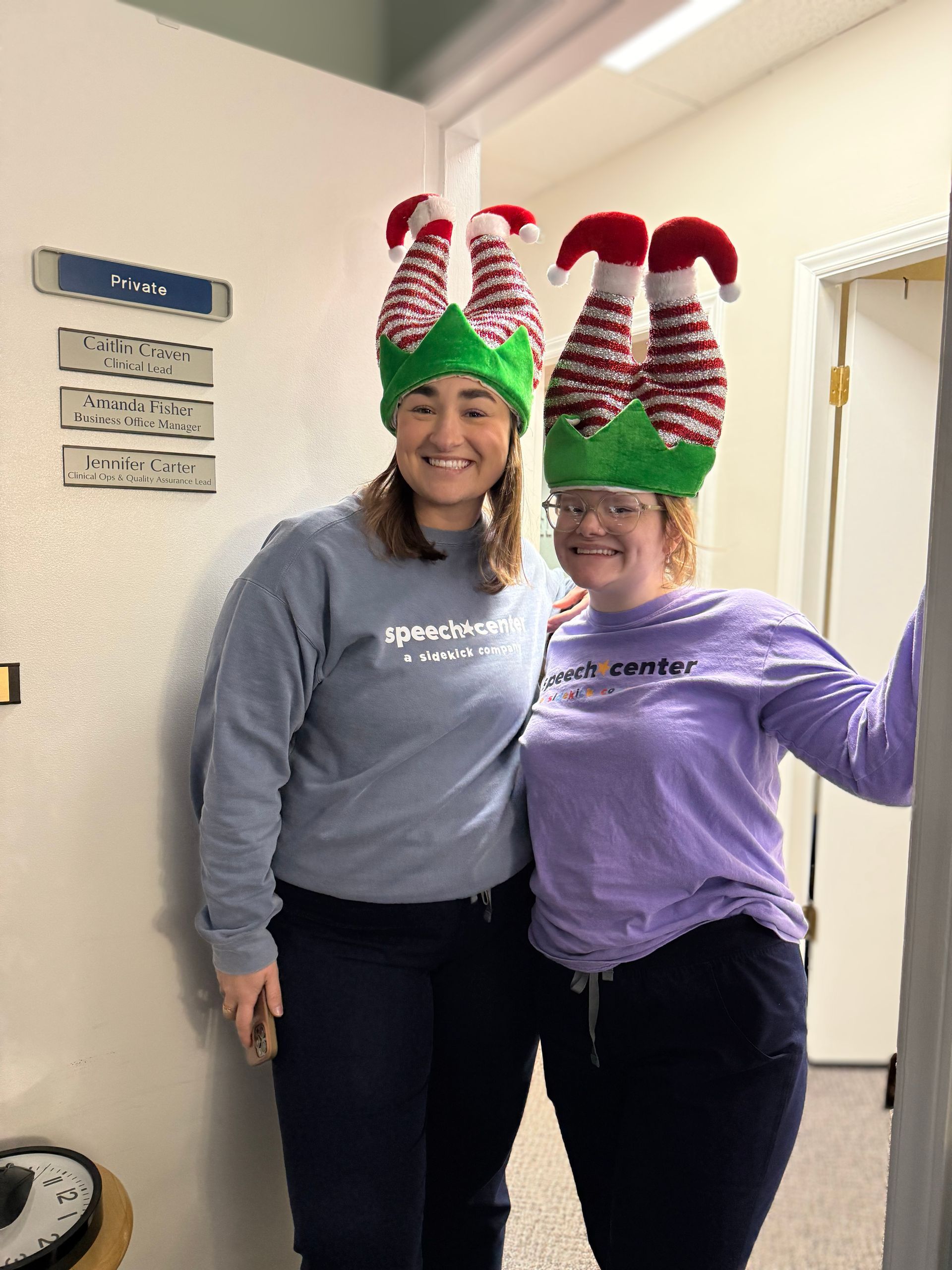 Two people wearing elf hats, smiling, in an office doorway. One in a blue sweatshirt, one in purple.