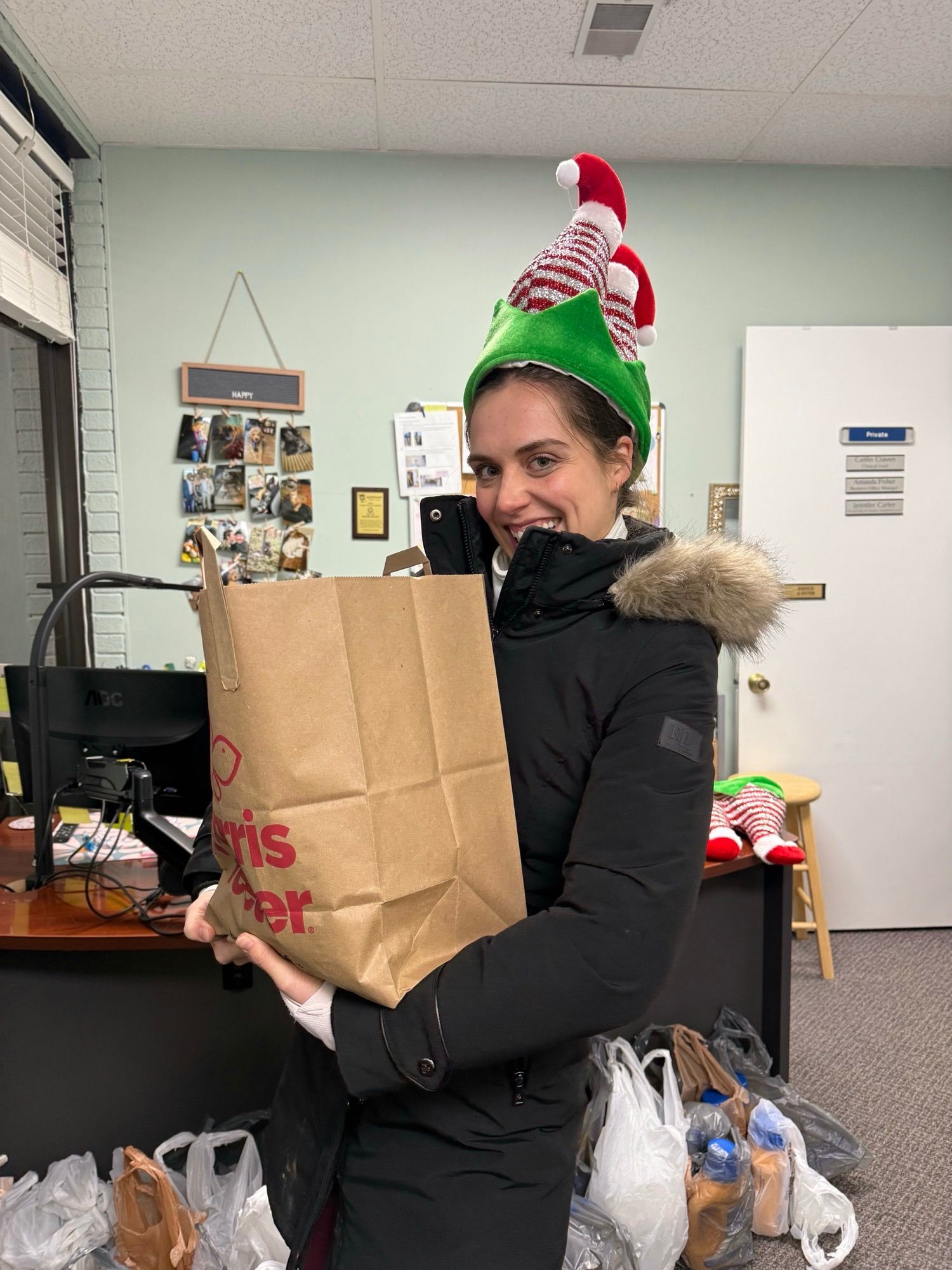 Woman in elf hat holding a grocery bag, smiling in an office.