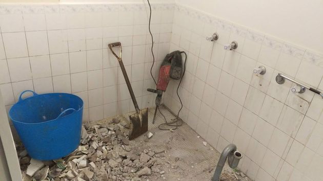 A bathroom undergoing renovation with tiled walls, debris on the floor, a blue bucket, a shovel, and a demolition hammer.