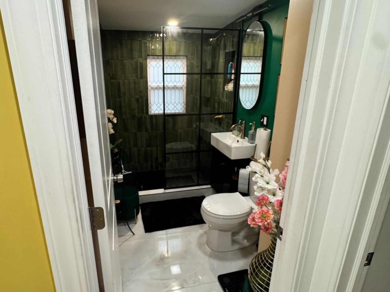 A modern bathroom with a glass-enclosed shower, green tiled walls, white floor tiles, a toilet, and a sink.