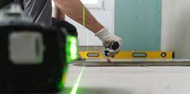 A worker in a glove uses a tool to level a floor, guided by a green laser line from a device in the foreground.