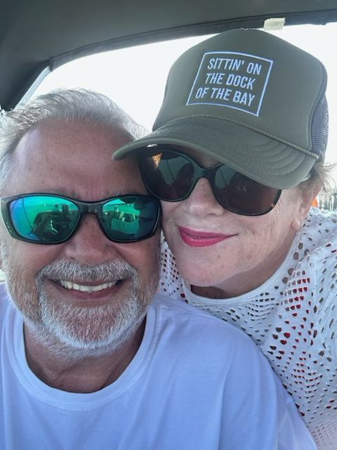 A man and a woman wearing sunglasses and a hat that says sitting on the dock of the bay
