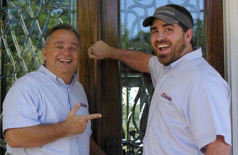 Two men are standing next to each other in front of a door and smiling.