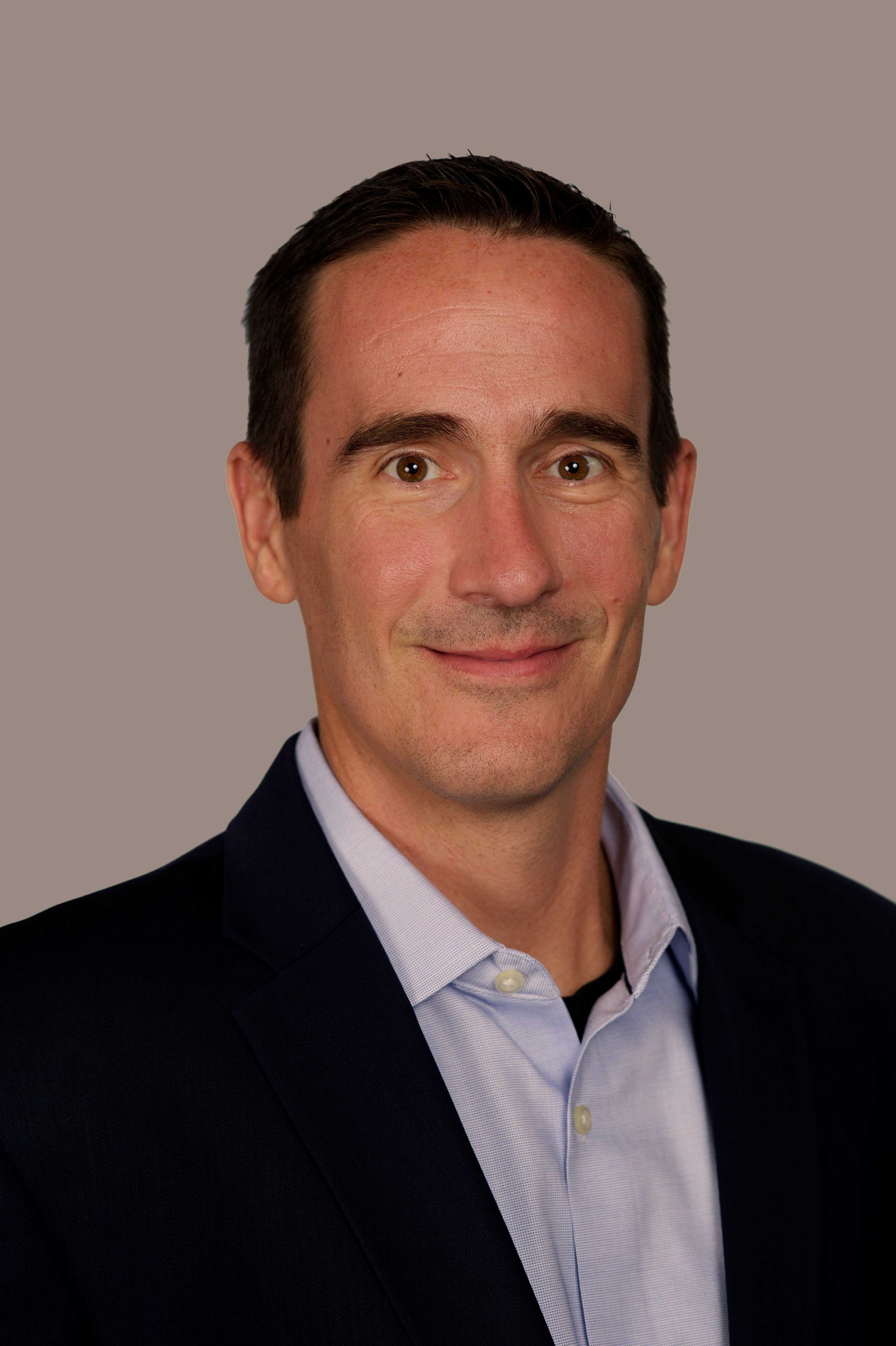 Man in a blazer and button-down shirt smiles, head and shoulders shot, against a gray background.