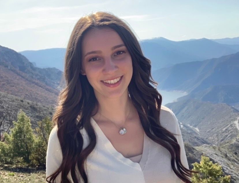 A woman in a white shirt is smiling in front of a mountain range