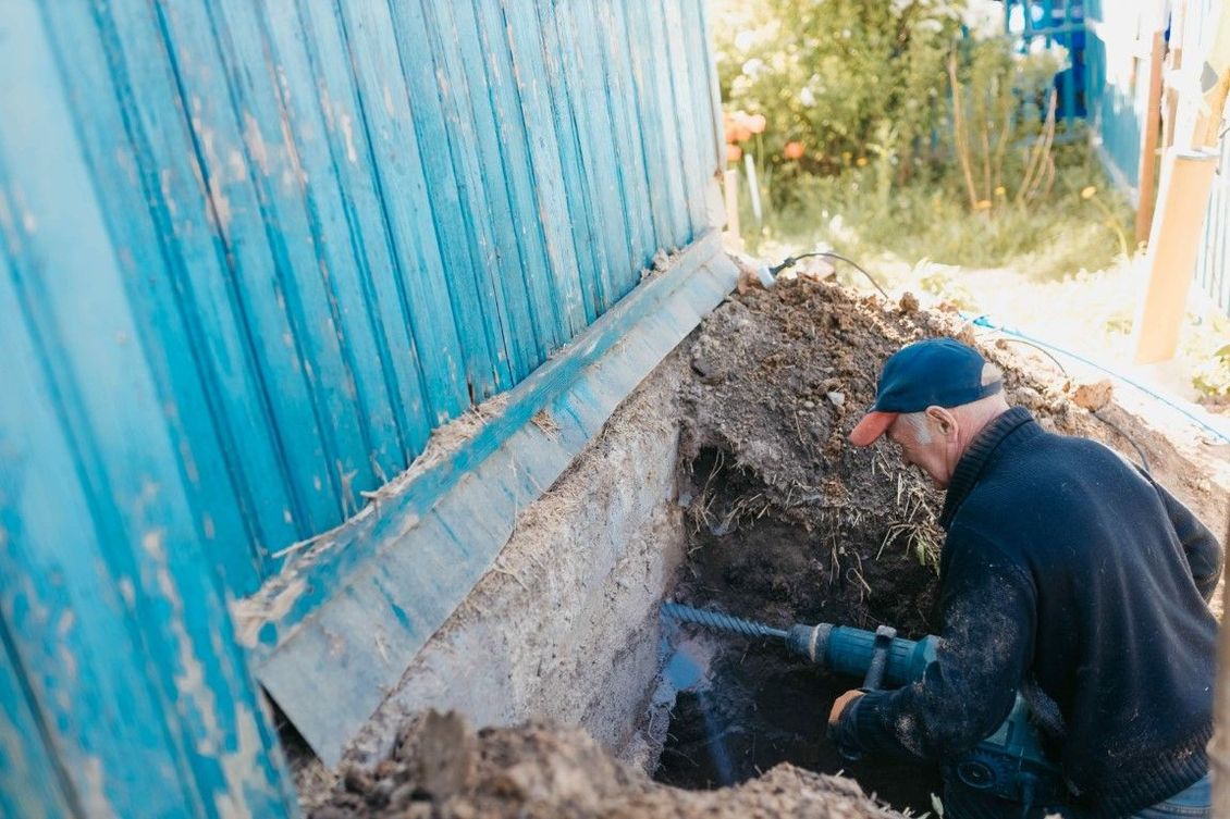 Man using a drill on a foundation next to a blue metal wall. Outdoors, daytime.
