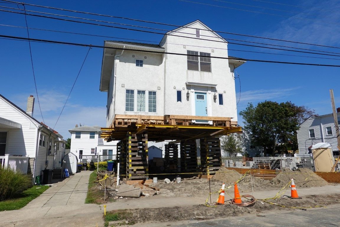 House lifted high on wooden supports, under construction on a sunny street with blue sky.