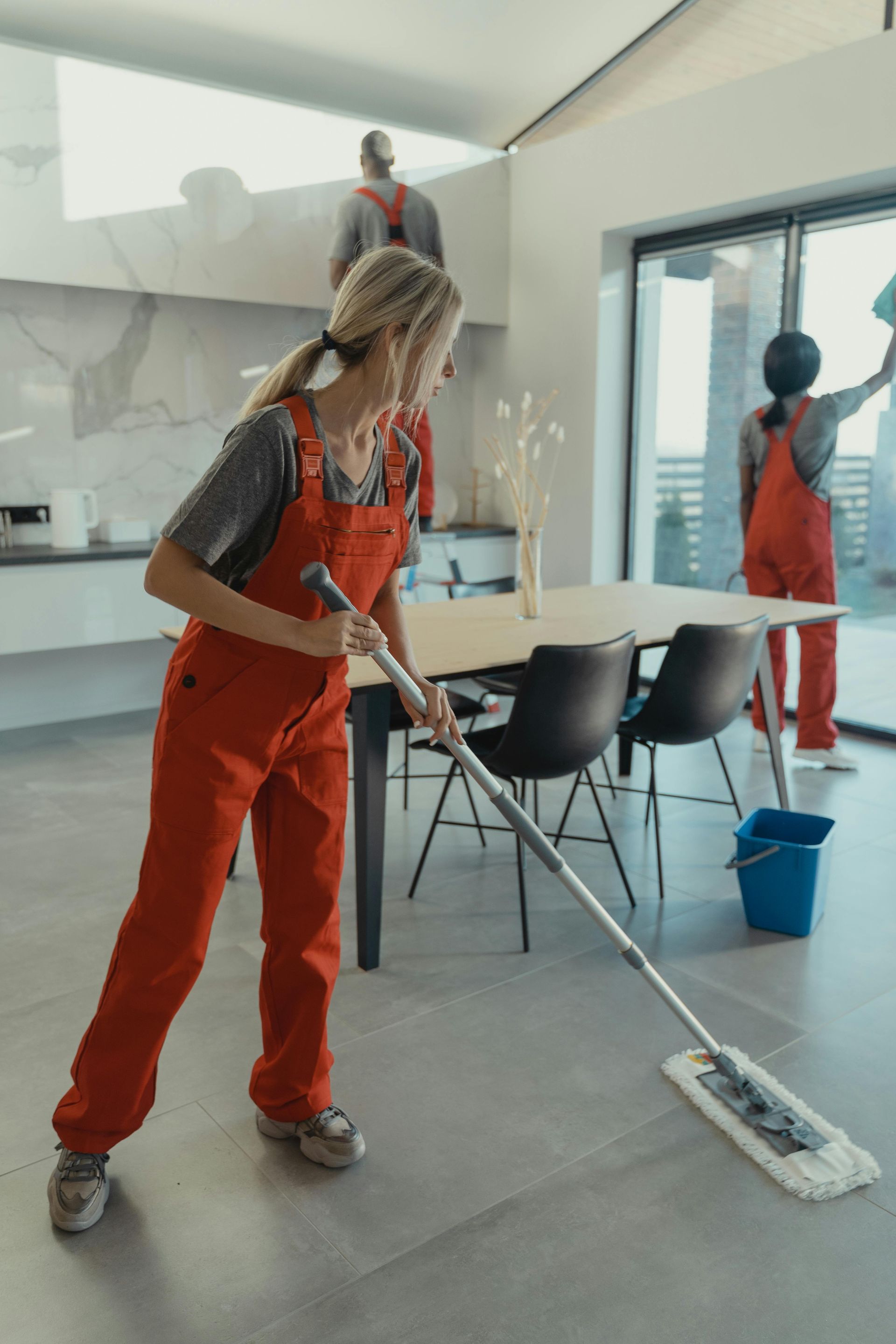Cleaning crew in orange jumpsuits cleaning a modern, sunlit room. One person mops the floor, another wipes a glass door.