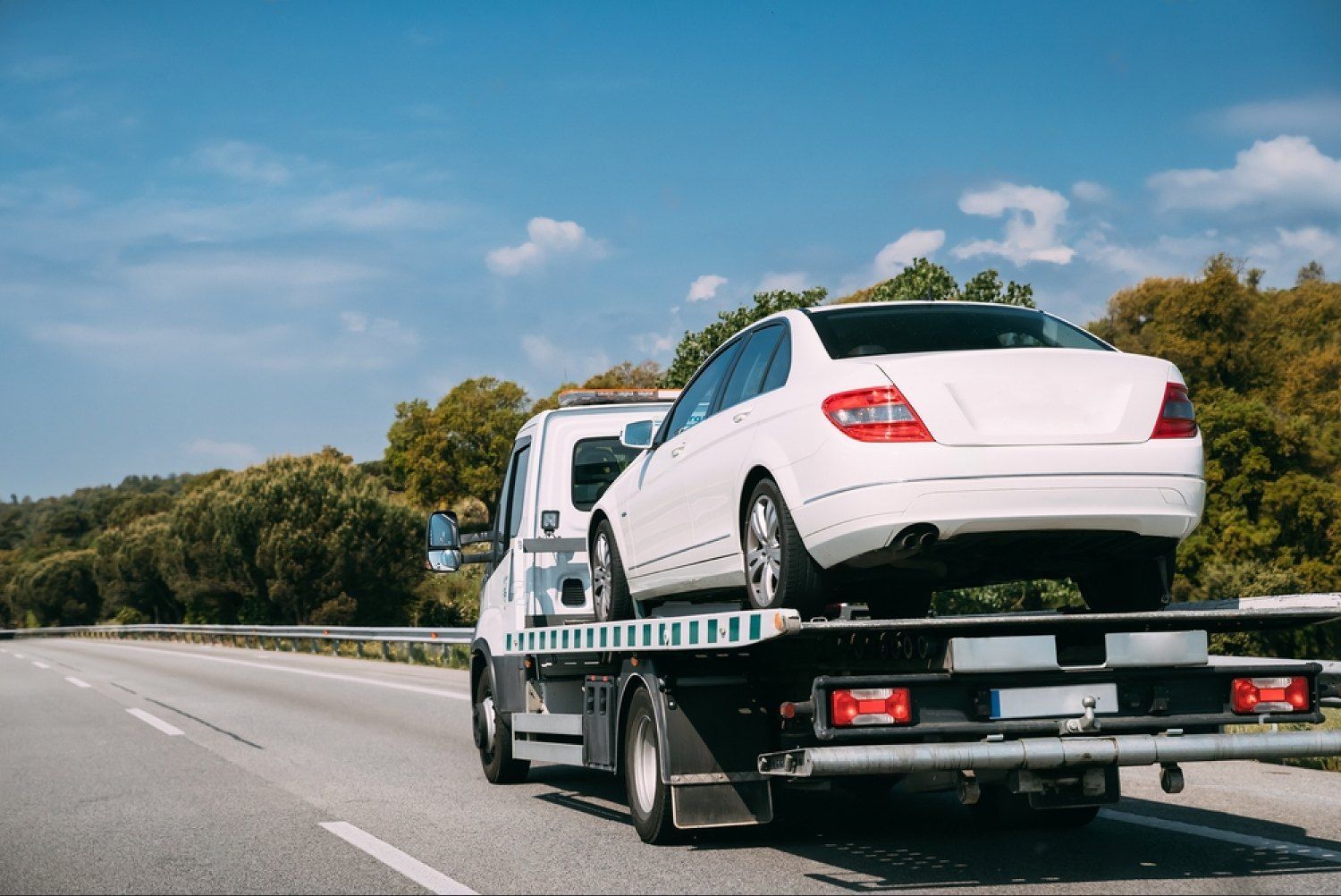 White car on a tow truck on a highway under a blue sky.