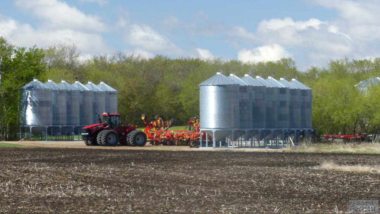 A Case-IH Steiger 4-wheel drive tractor with an air seeder is parked between rows of grain bins next to a cultivated field.
