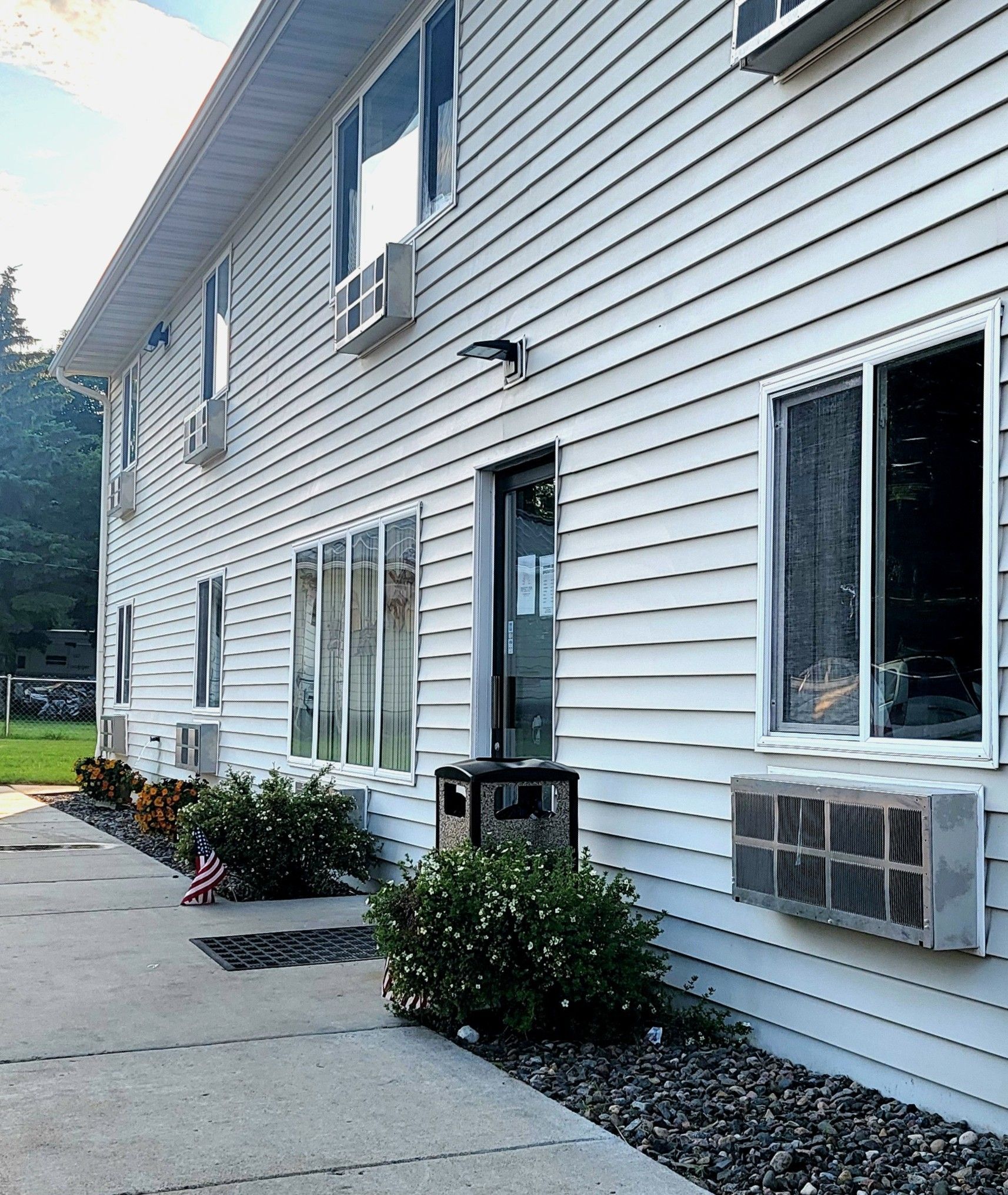 White building with multiple windows, air conditioners, and a door, next to a sidewalk and bushes.