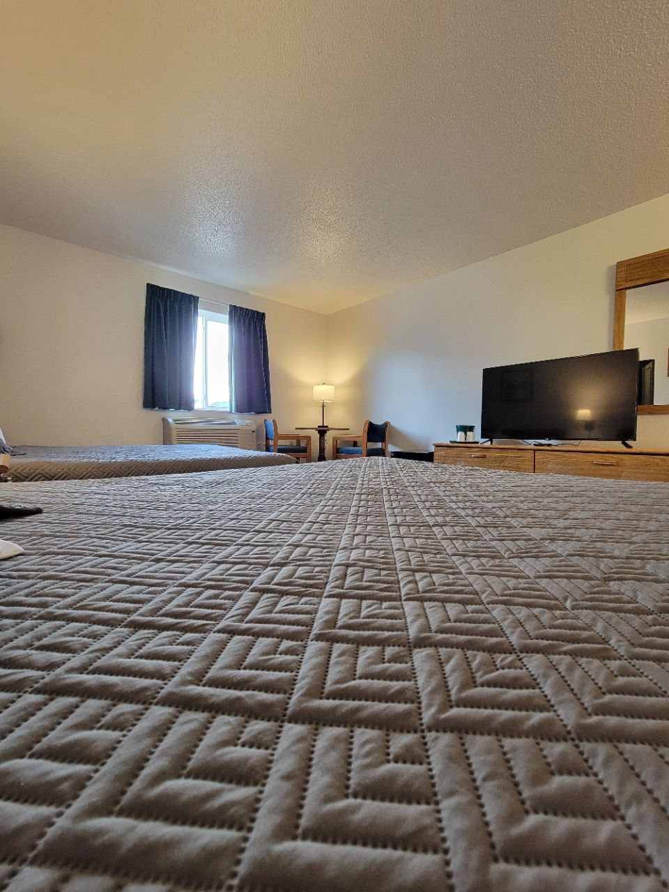 Bedroom with two beds, television, window, and a lamp. Walls are white and the carpet is light gray.