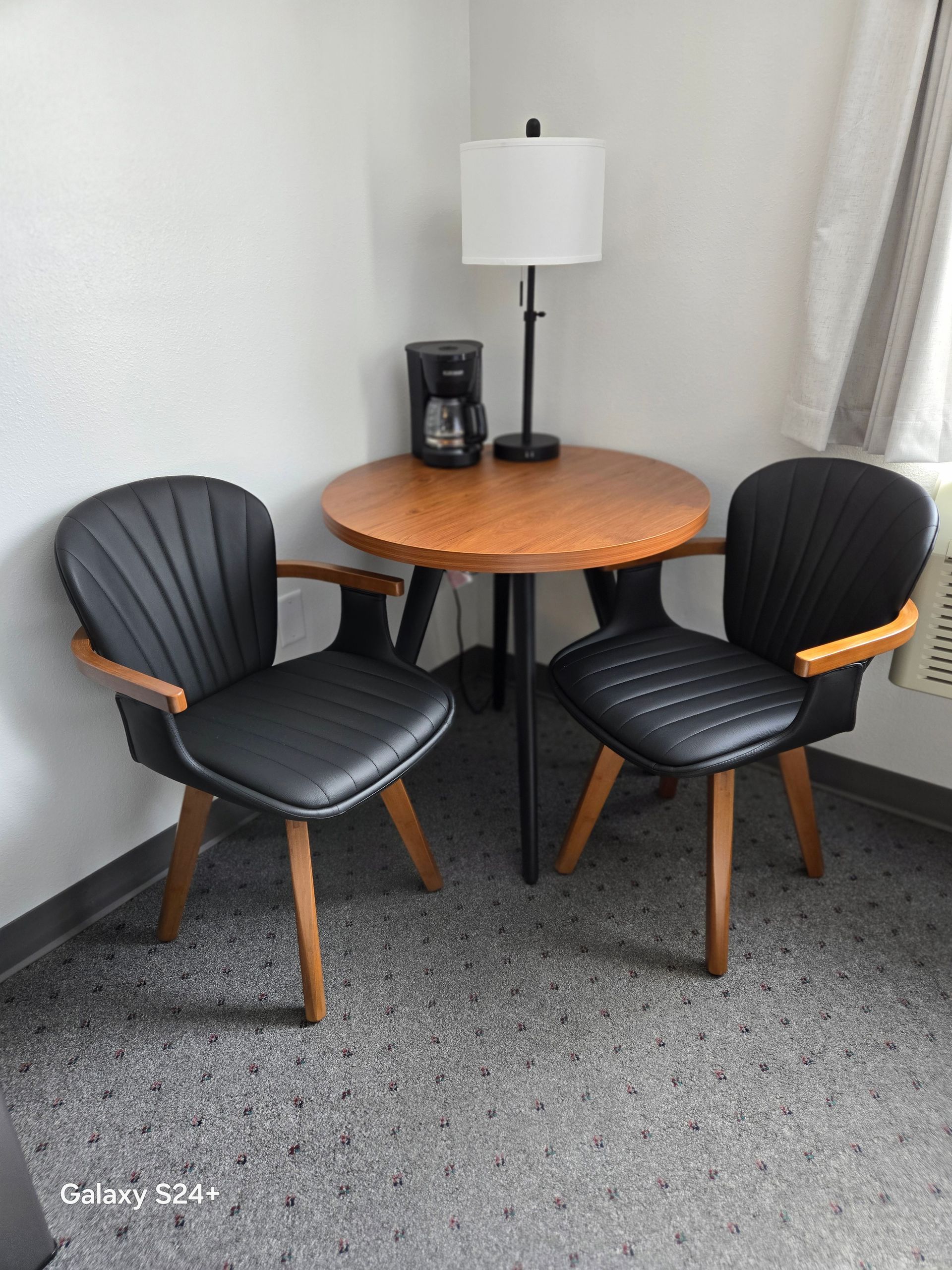 A round table with a lamp and coffee maker, flanked by two black leather chairs, set against a white wall.