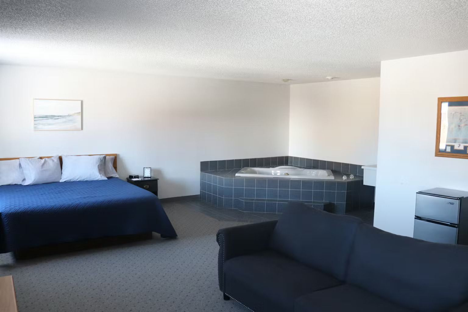 Hotel room with a bed, hot tub, and small refrigerator. Blue and white accents on neutral walls.