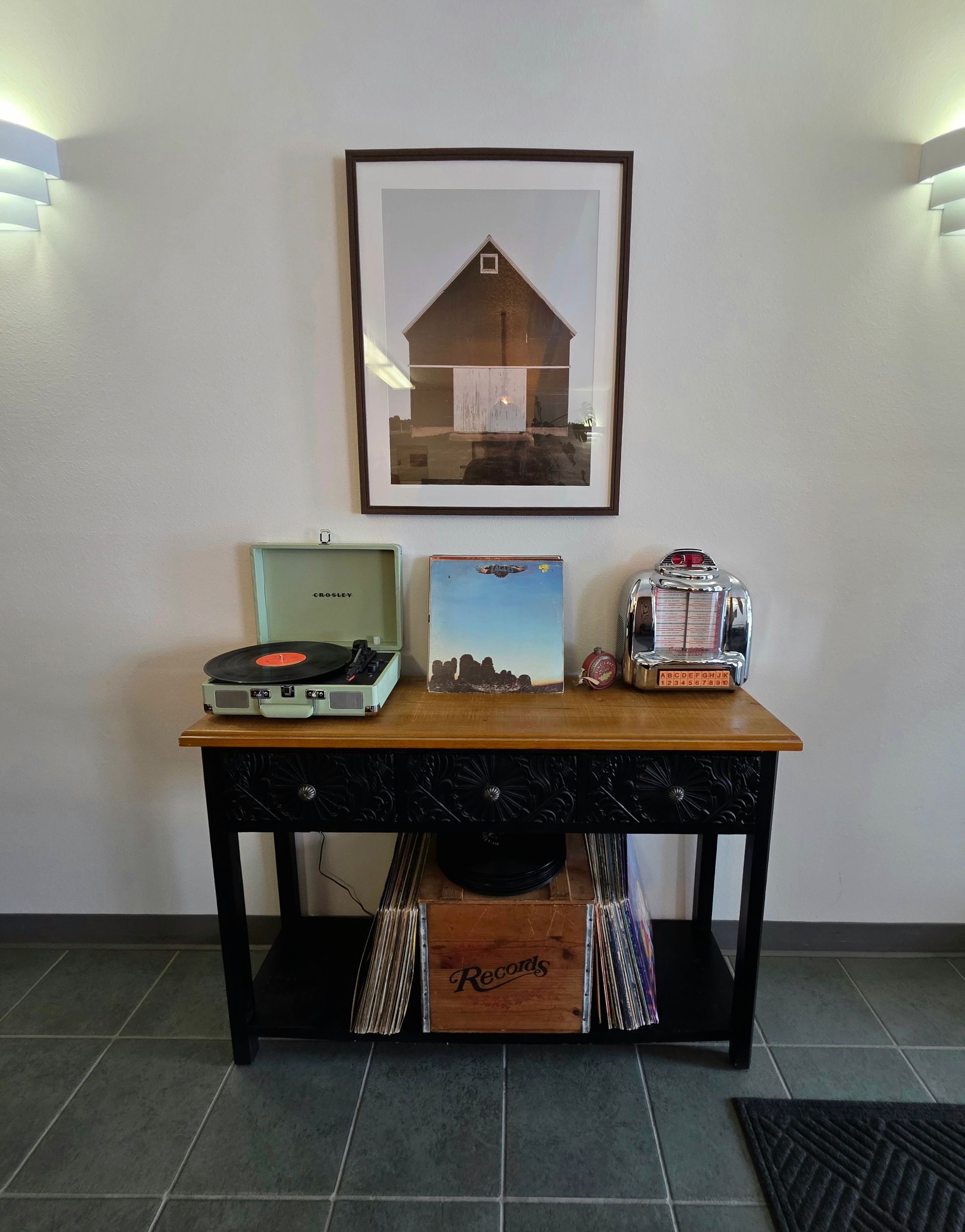 A wooden table with a record player, records, and a jukebox below a framed barn photo.