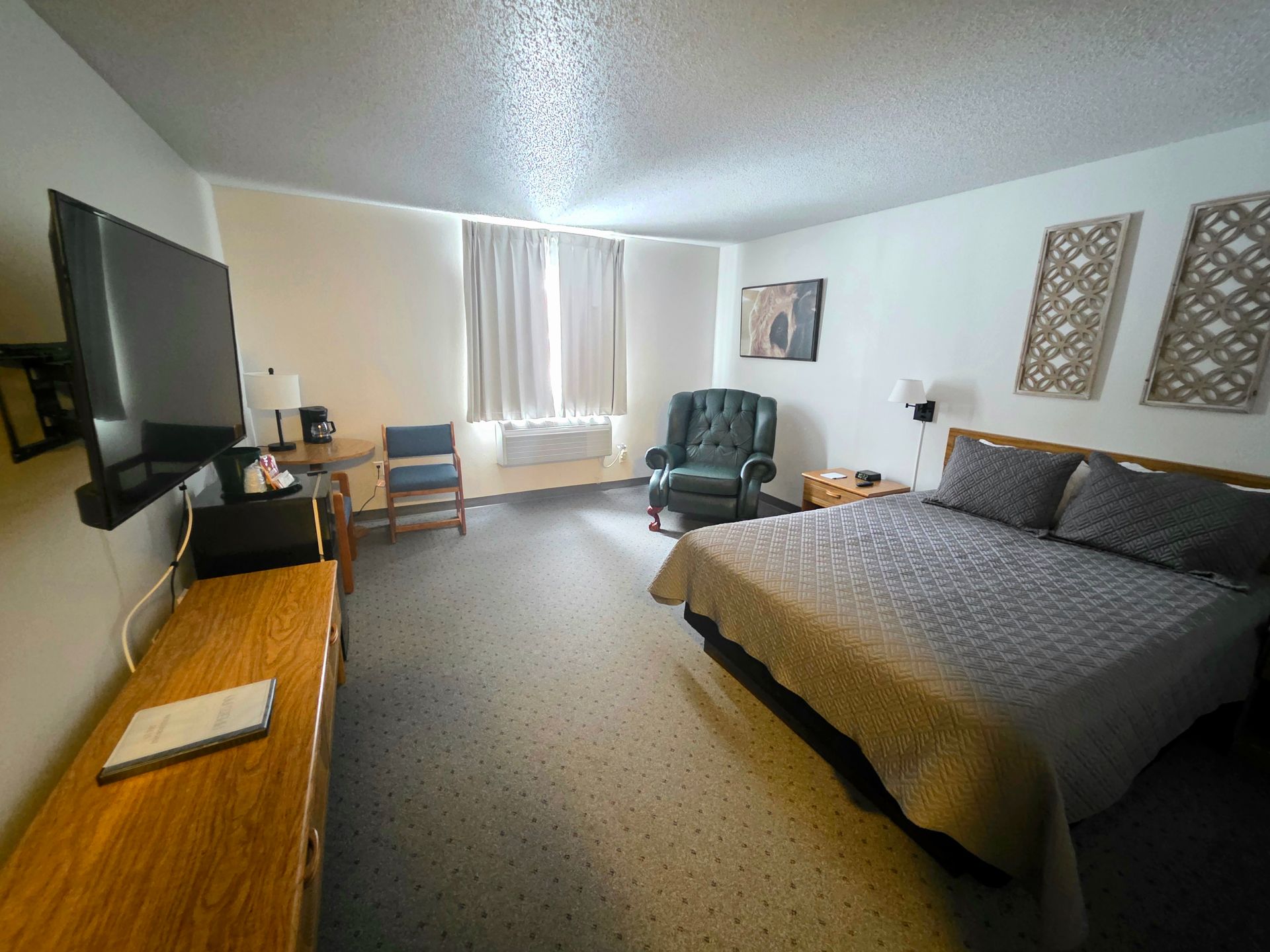 Hotel room interior: bed, armchair, TV, desk, and round table near window.