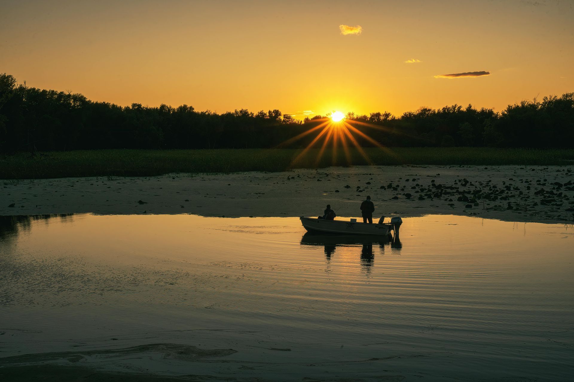 Two people fishing from a boat on a lake at sunset. Silhouette of trees and sunburst in the orange sky.