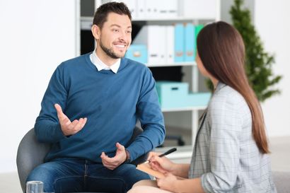 Man smiling, gesturing, speaking to person taking notes in a professional office setting.