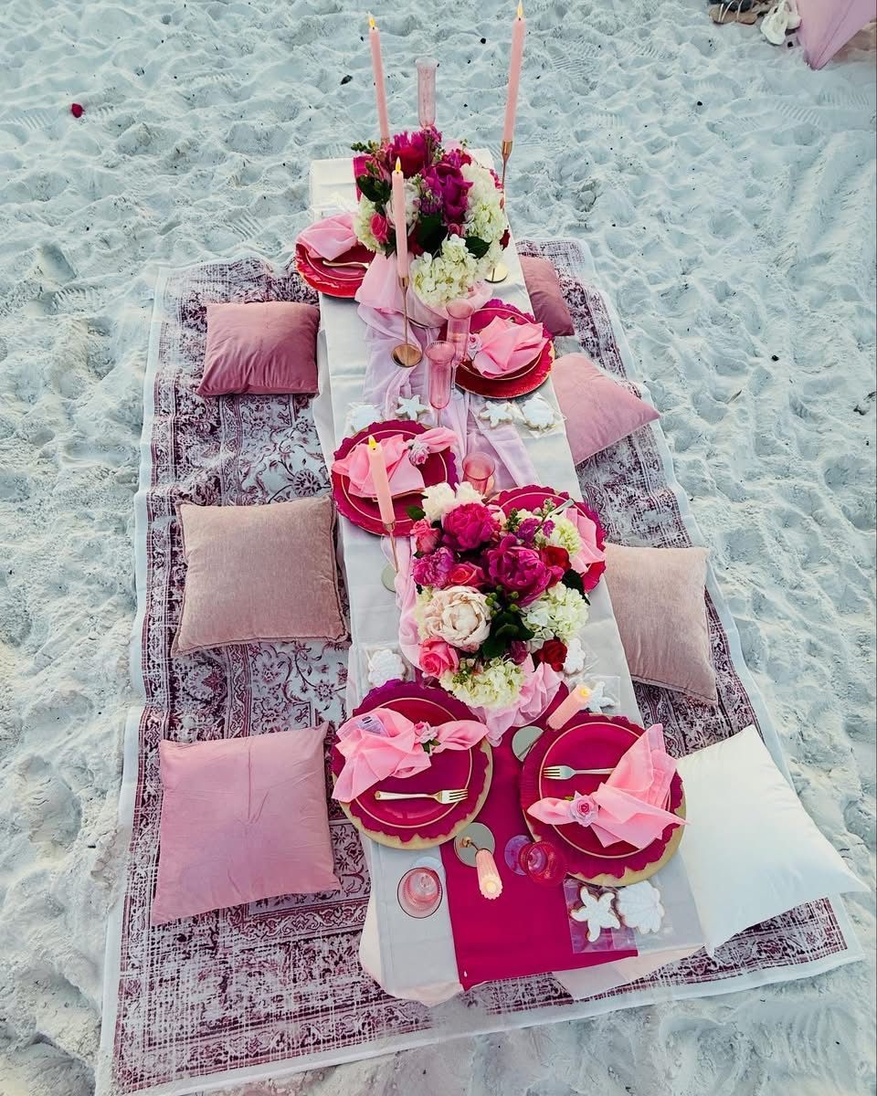 Picnic setup on a beach: flowers, pink dishes, pillows, candles on a blanket.