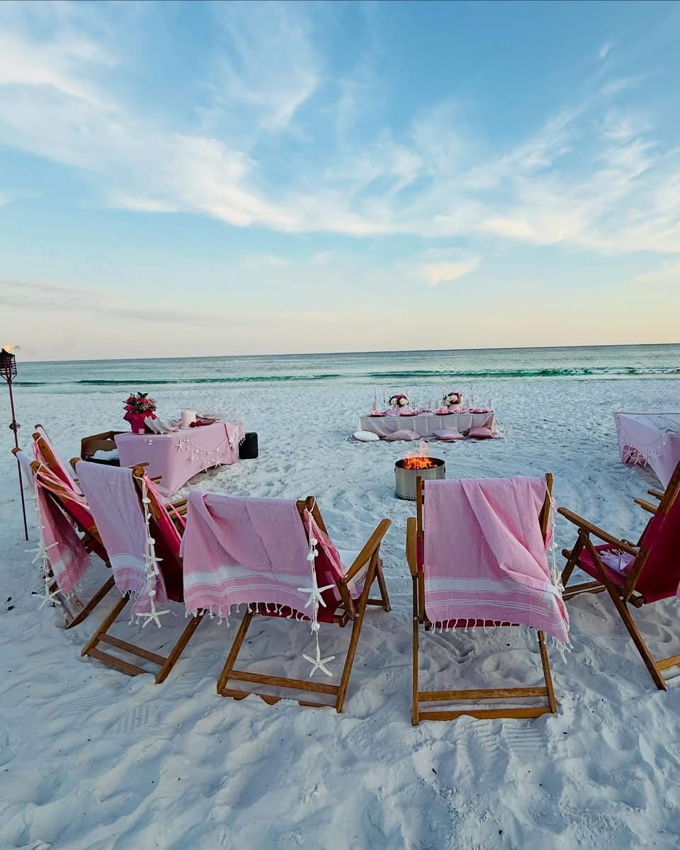 Beach picnic setup with pink towels, chairs, and tables on white sand.