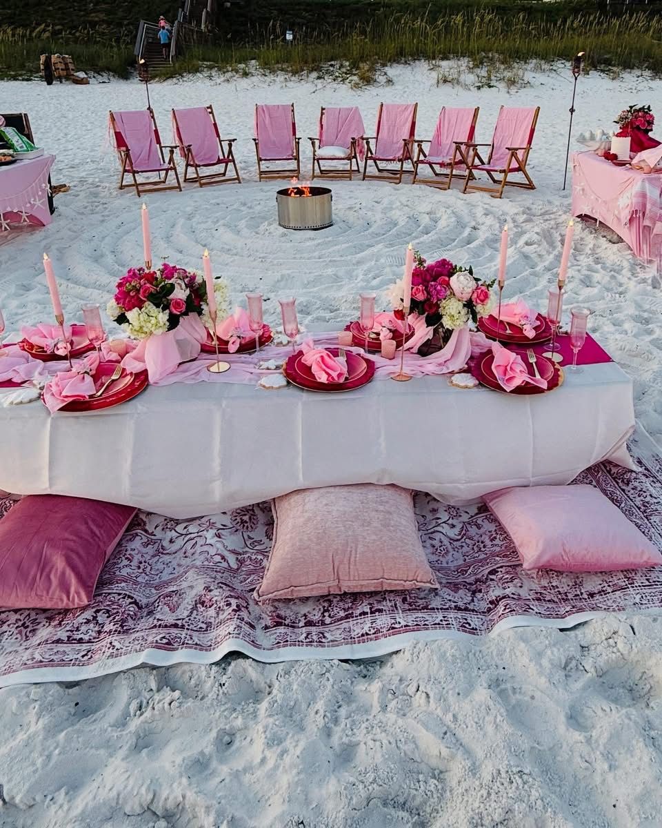 Beach picnic setup: pink decor, low table, seating, fire pit, flowers, soft light, sandy shore.