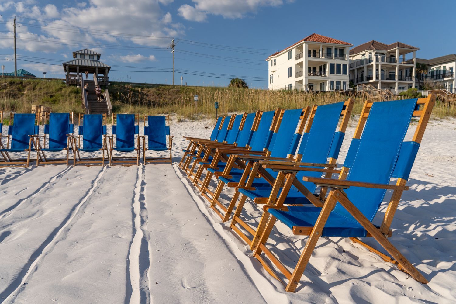 Blue beach chairs in rows on white sand, with a multi-story building and a gazebo in the background.