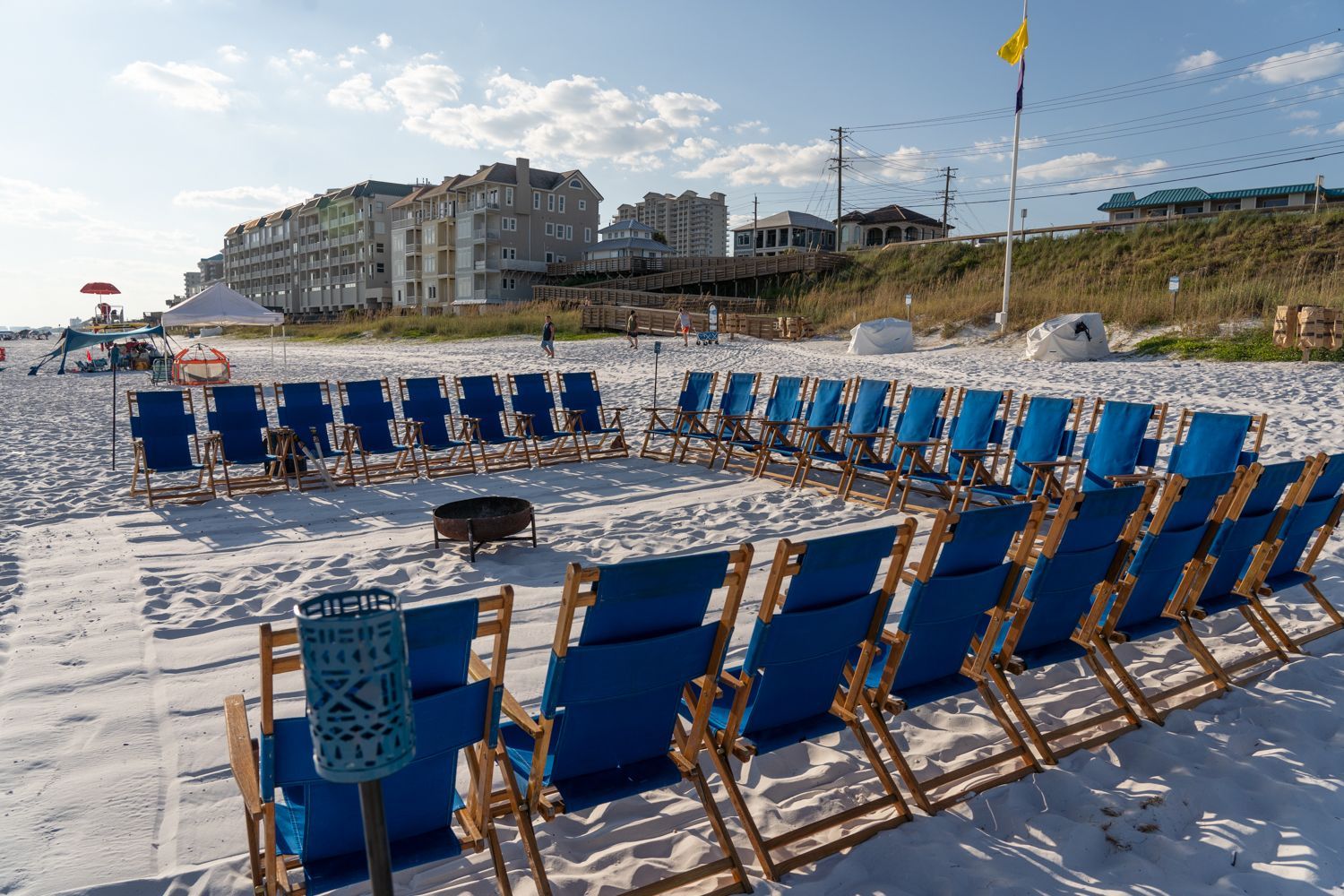 Blue beach chairs arranged in a circle on sand, fire pit, buildings in the background.