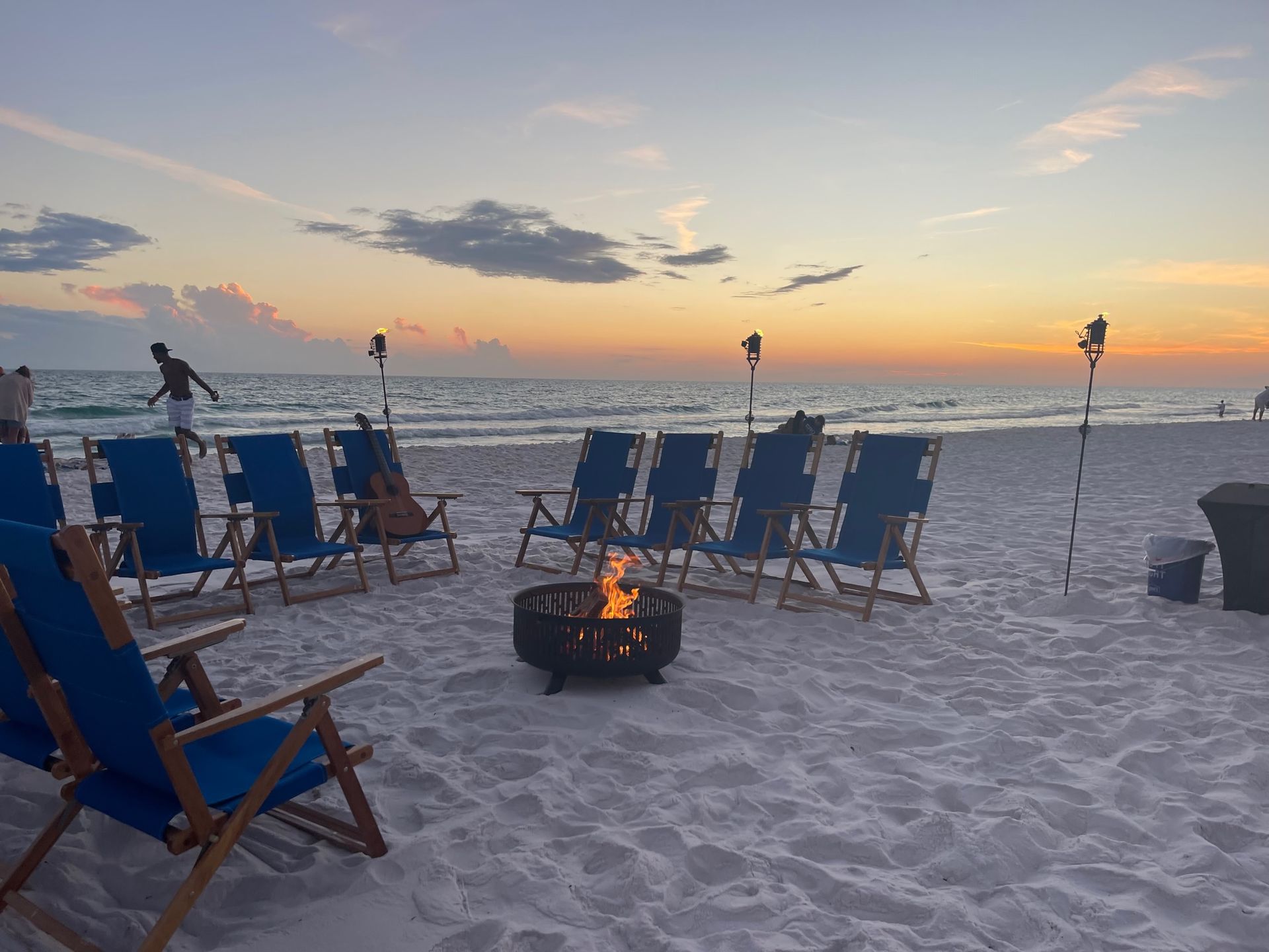 Beach scene at sunset with a fire pit, blue chairs, and tiki torches.