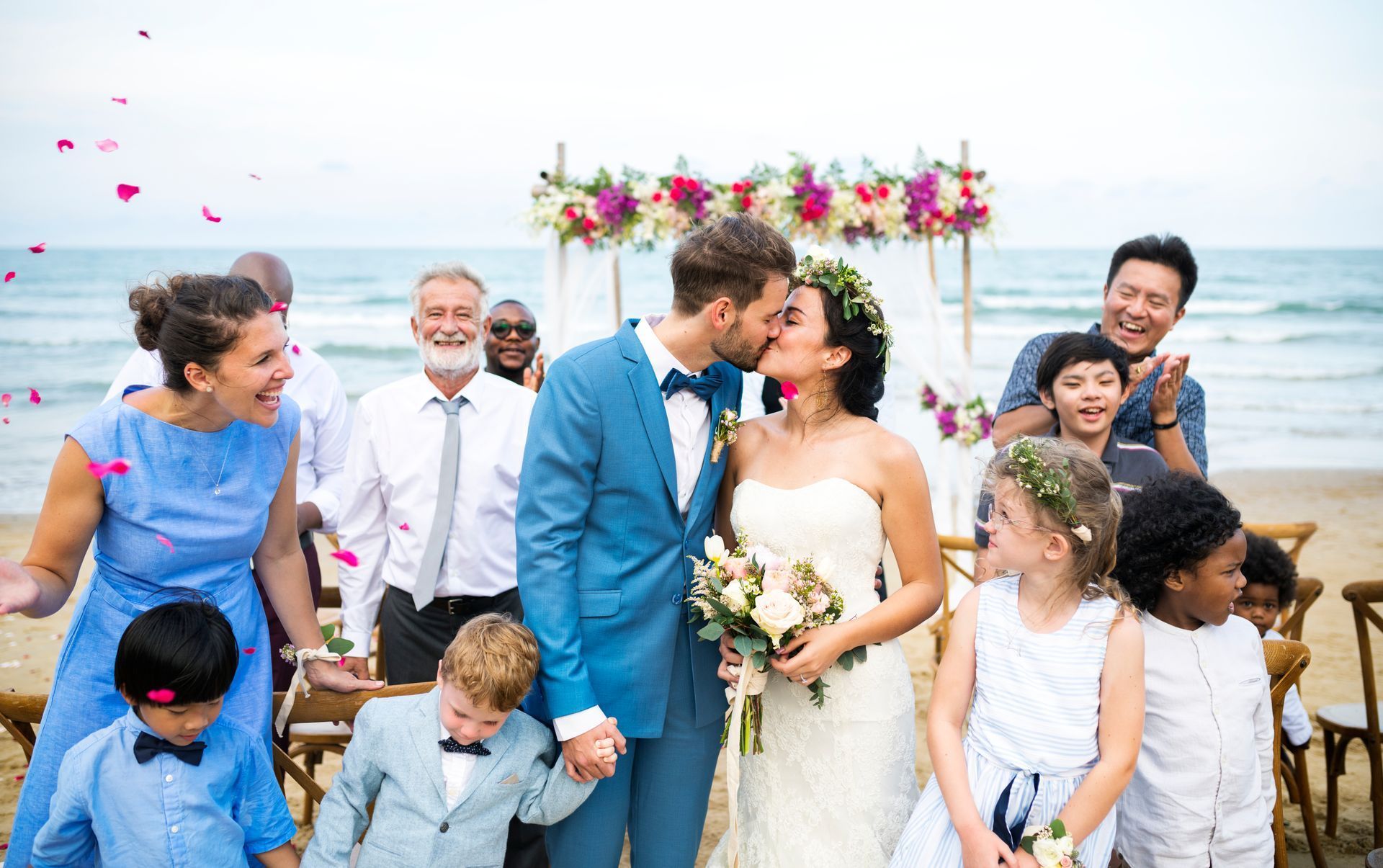 A bride and groom kissing in front of their wedding party on the beach.