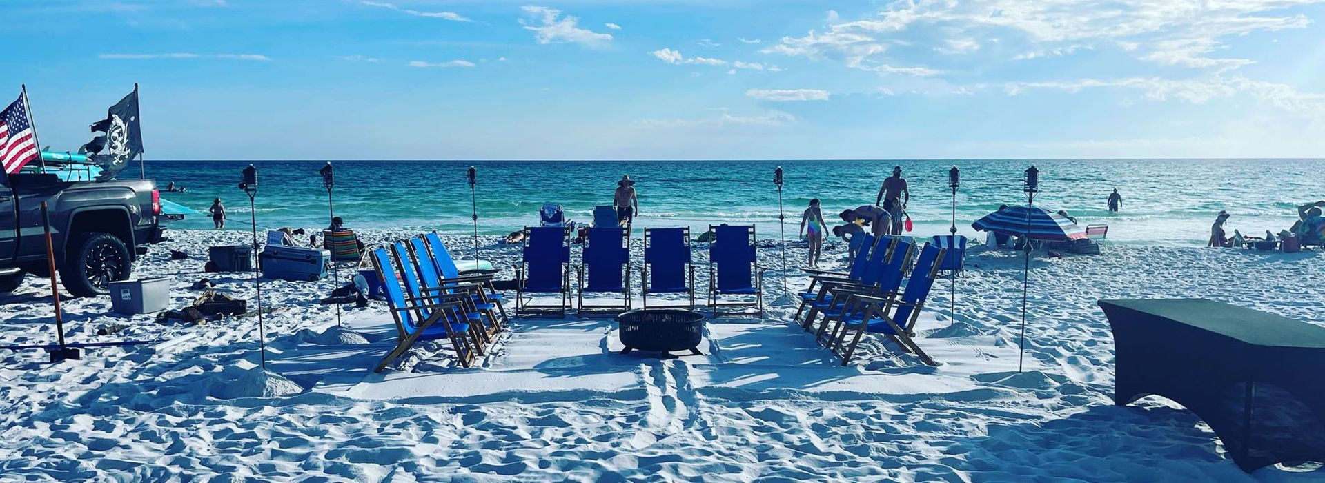 Beach scene with blue chairs, fire pit, and people enjoying the ocean. American flag flies.