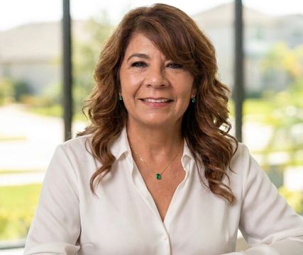 Smiling woman in a white blouse, seated indoors with a bright windowed background.