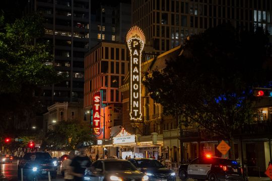 Nighttime city street with a lit Capitol Theatre marquee, traffic, and tall buildings in the background