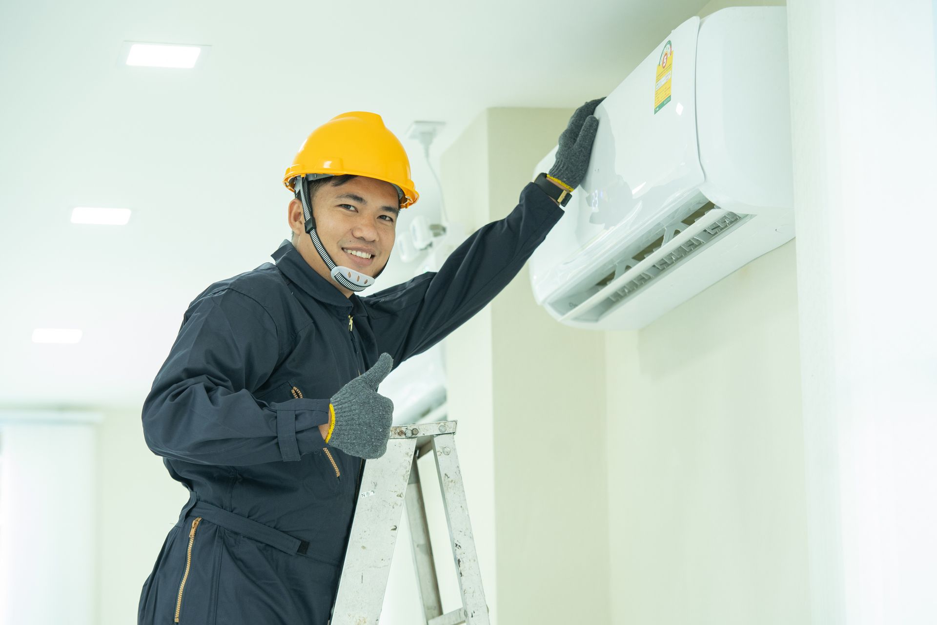 A man in a hard hat and overalls stands on an upstart after air conditioning installation.