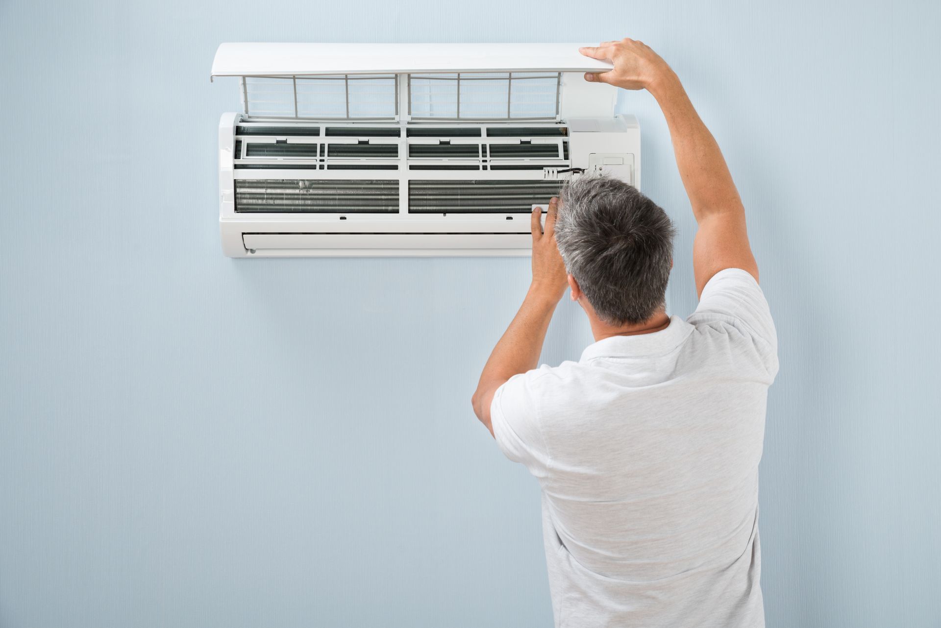 Rear view of a man wearing a white shirt, opening an air conditioner and checking its filters.