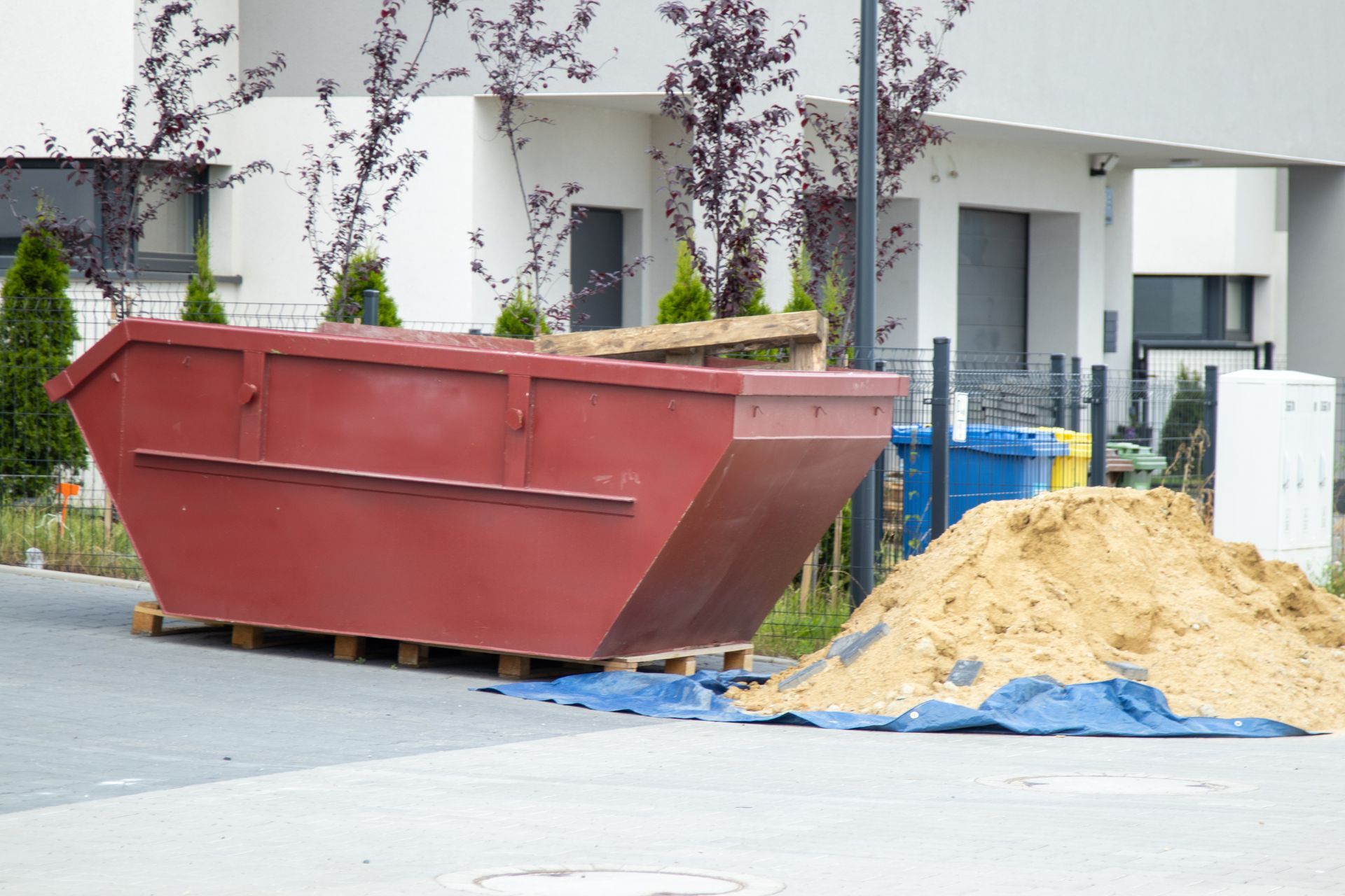 Red dumpster on a pallet, beside a pile of sand, in front of a white building.
