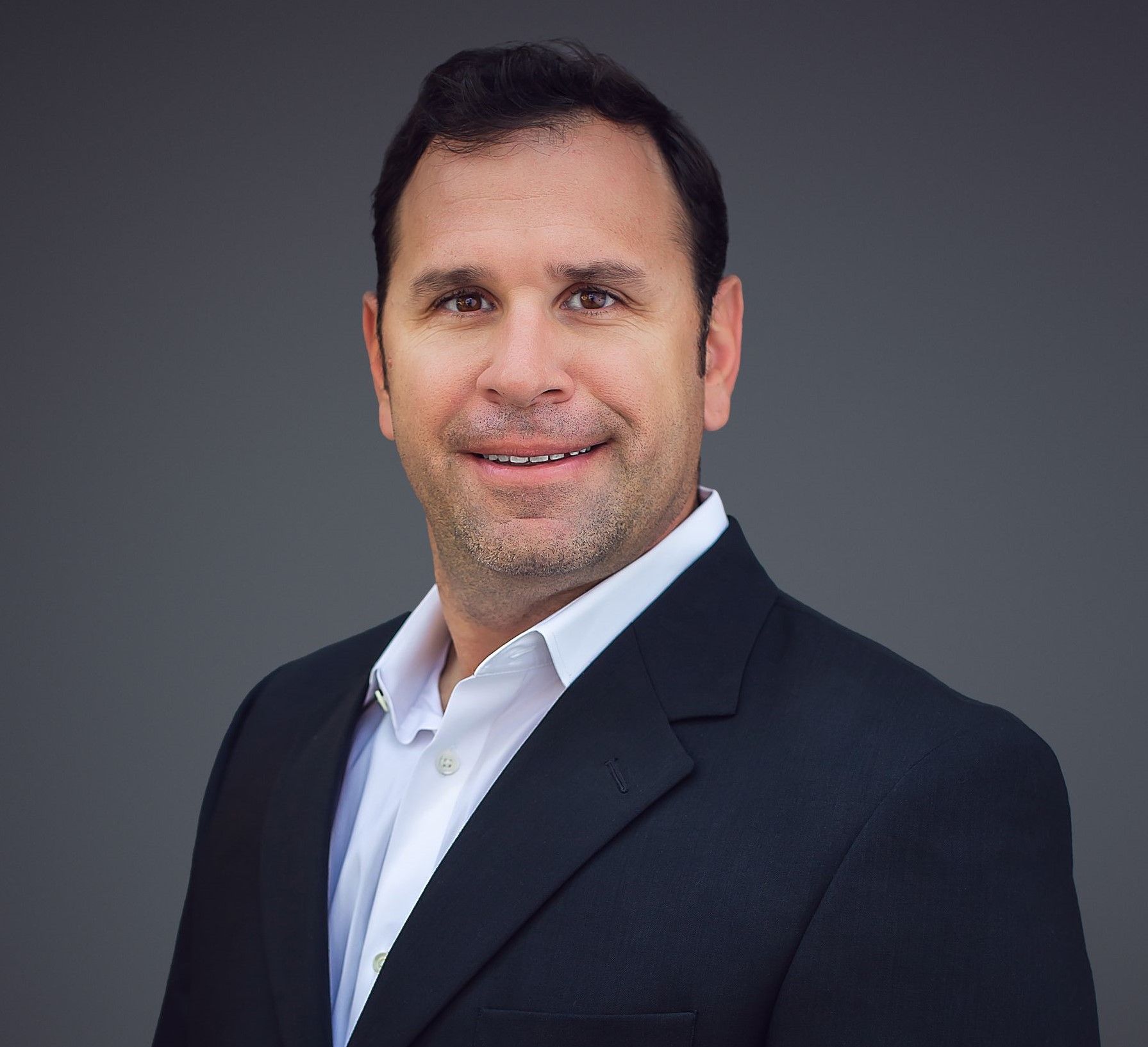 Man in a blazer and open-collared shirt smiling at the camera against a gray background.