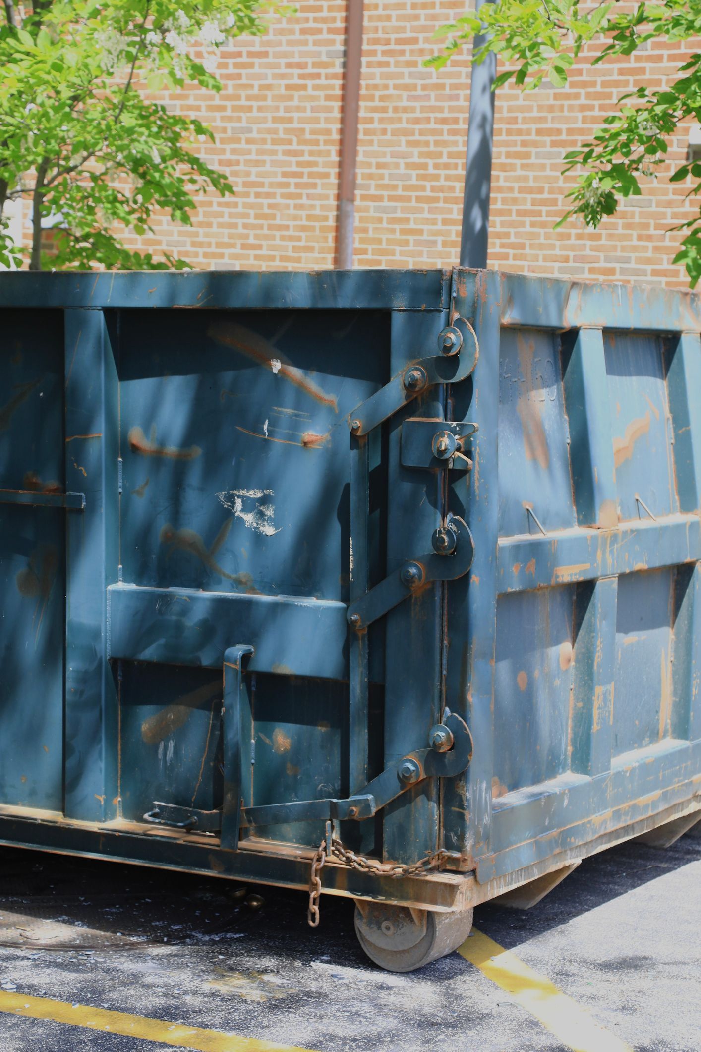 Blue dumpster with wheels, side panel with hinges and latch, set against a brick building.