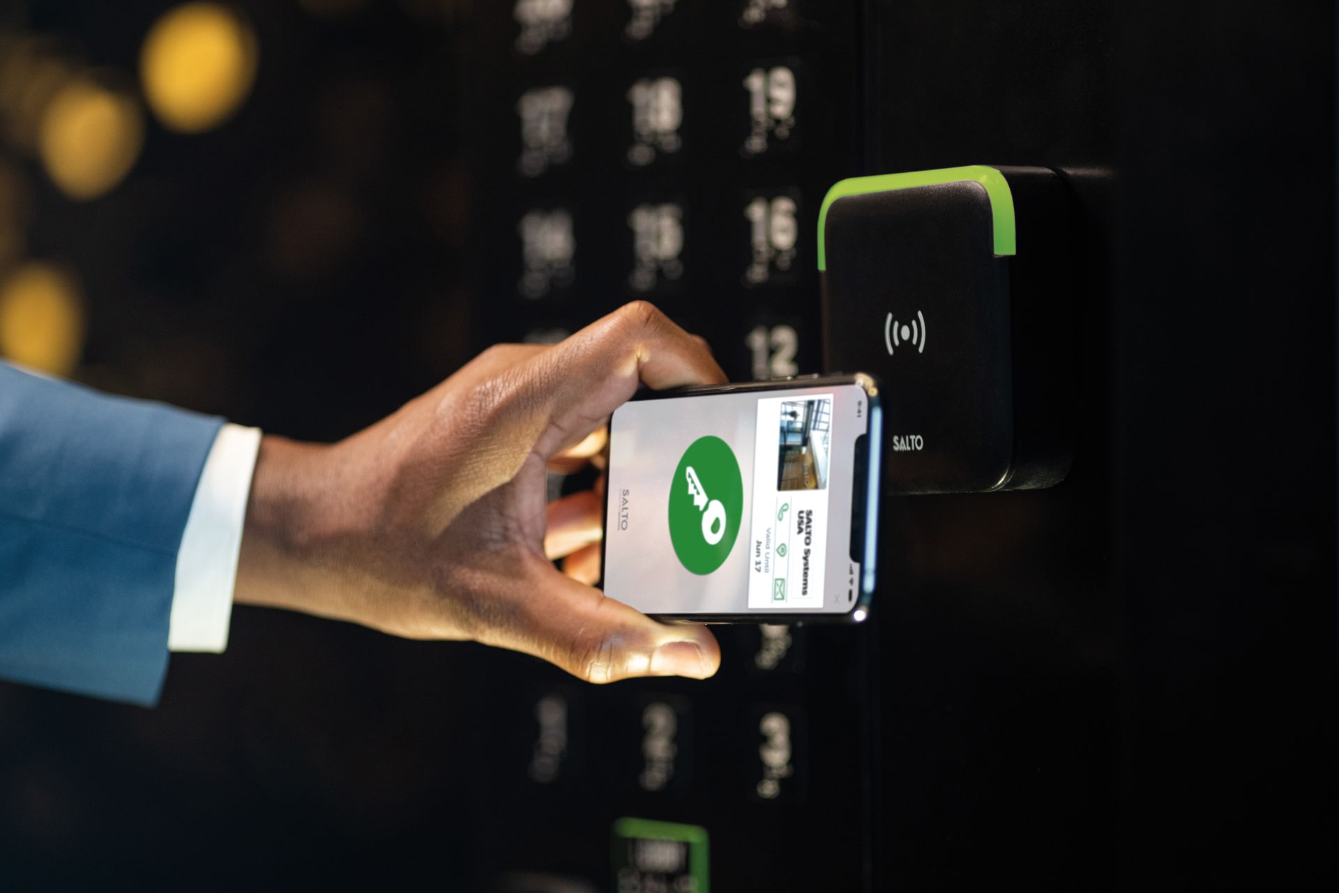 A hand holds a smartphone displaying a green key icon near an NFC contactless reader on a dark panel.