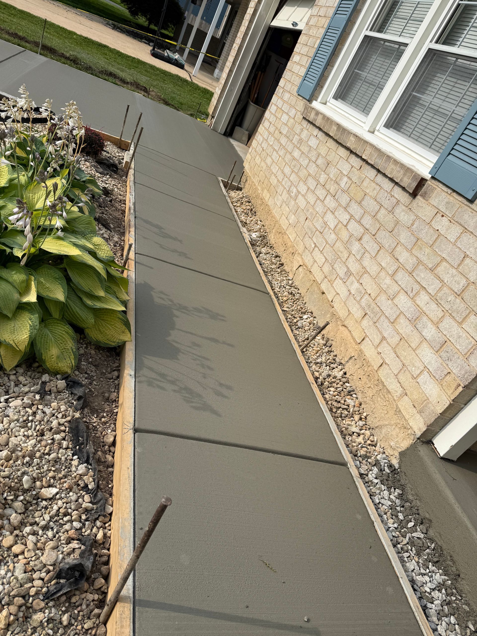 Freshly poured concrete sidewalk next to a house with blue shutters and a small gravel bed.
