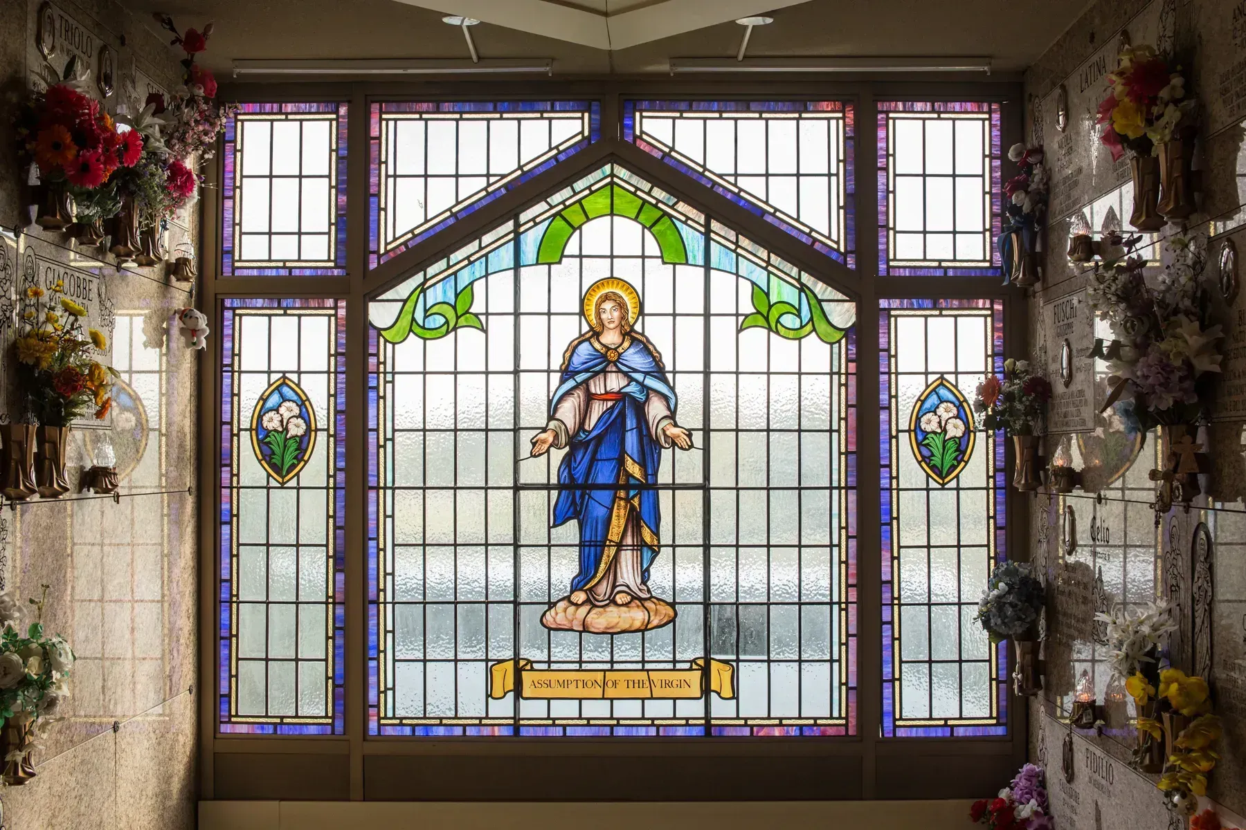 Stained-glass window of the Virgin Mary in blue robes, surrounded by floral decorations.