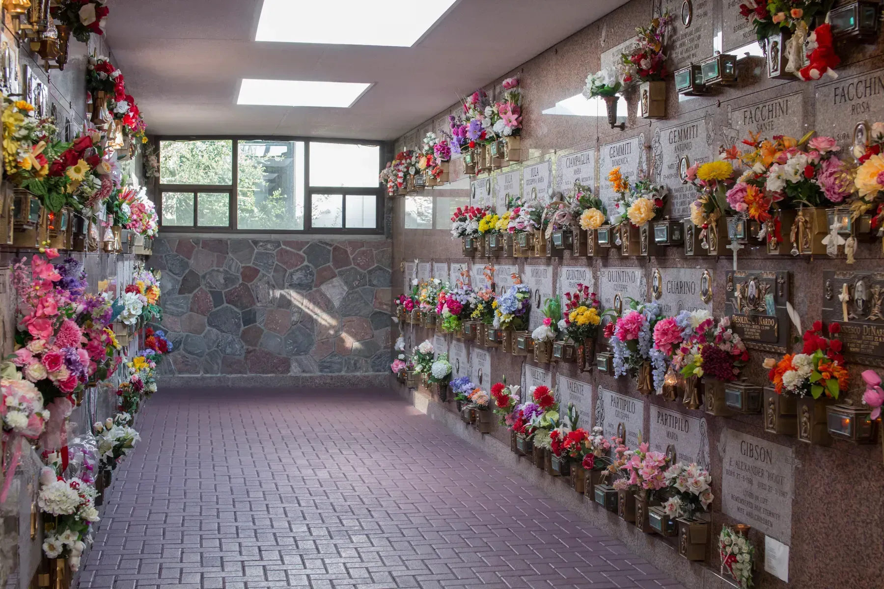 Flower-covered memorial wall in a brick corridor with a distant mural and skylights