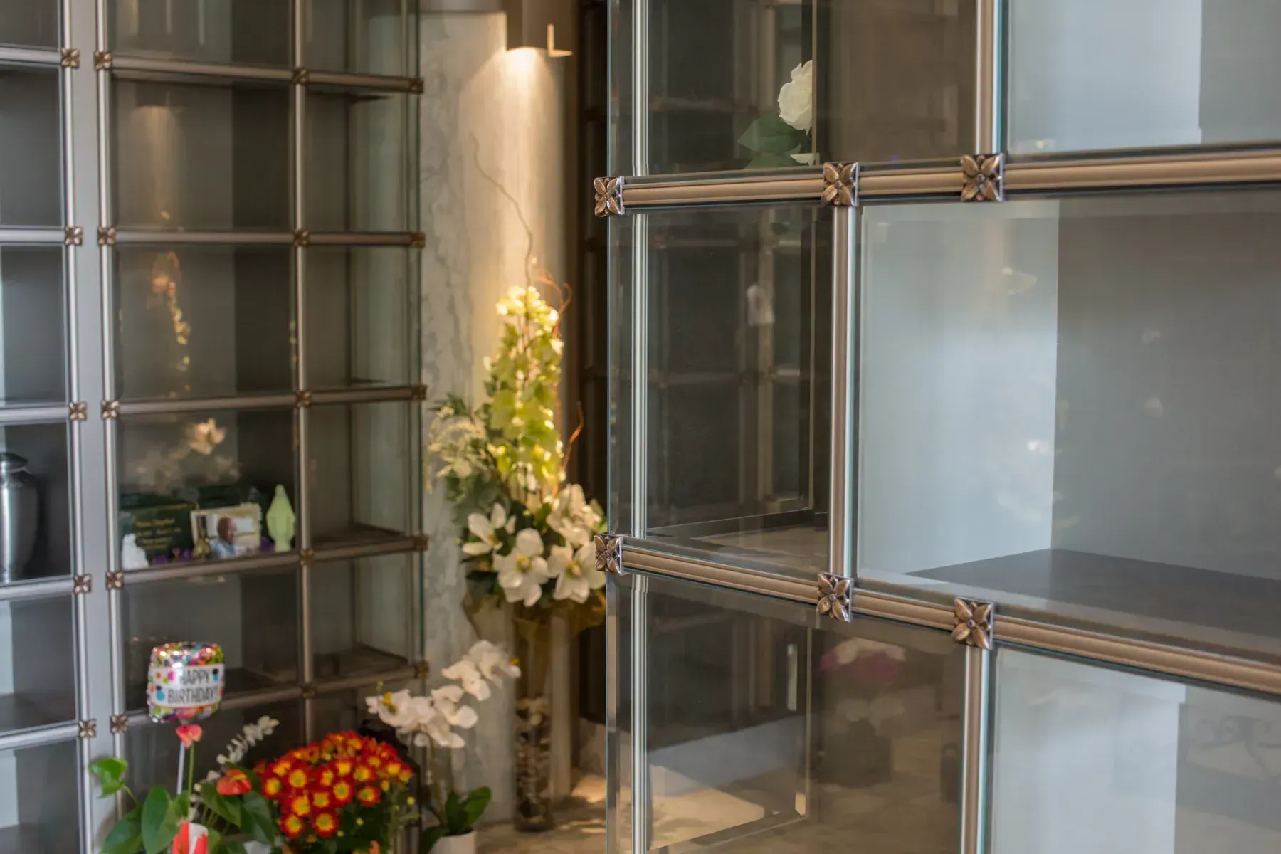 Glass memorial wall with shelves, flowers, and a wreath in a lobby or mausoleum