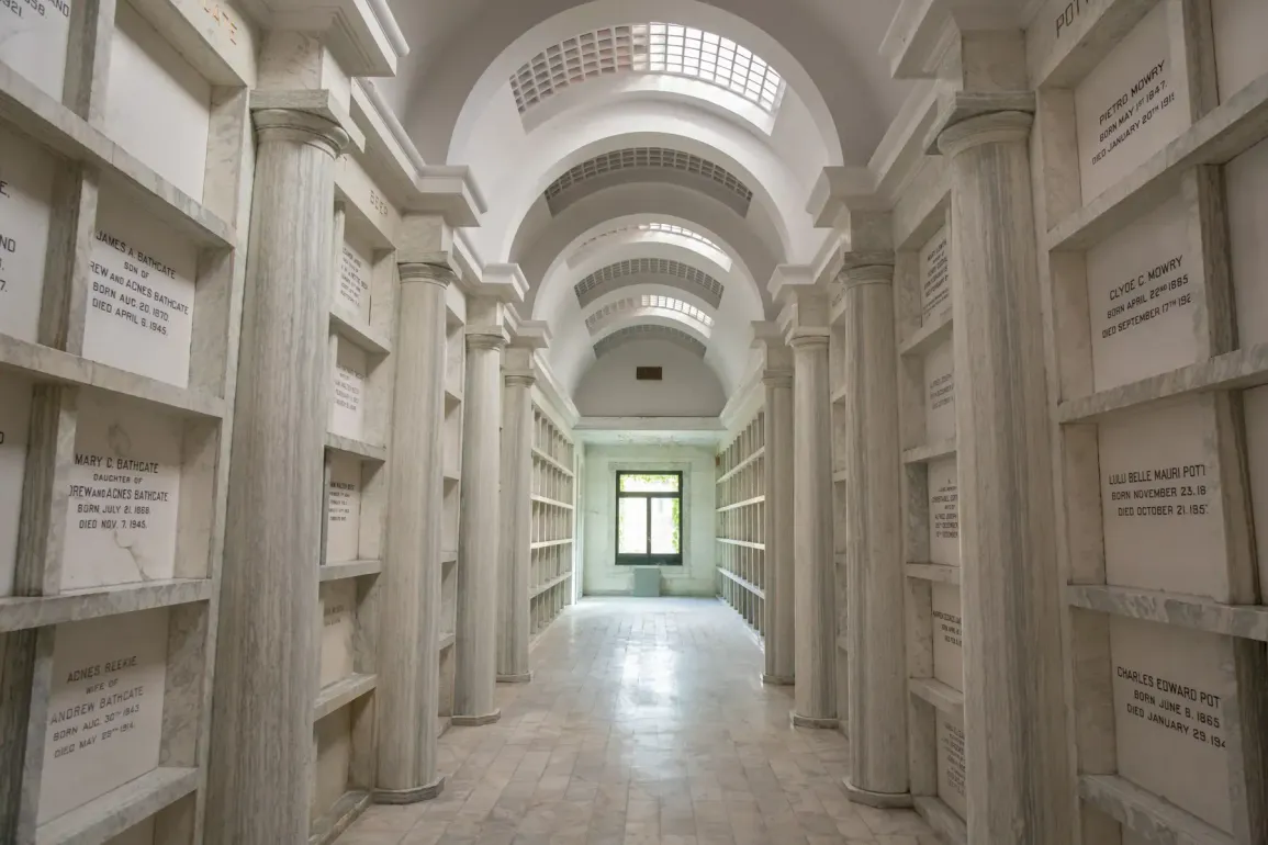 White mausoleum corridor with arched ceiling, stone niches, and a bright doorway at the end