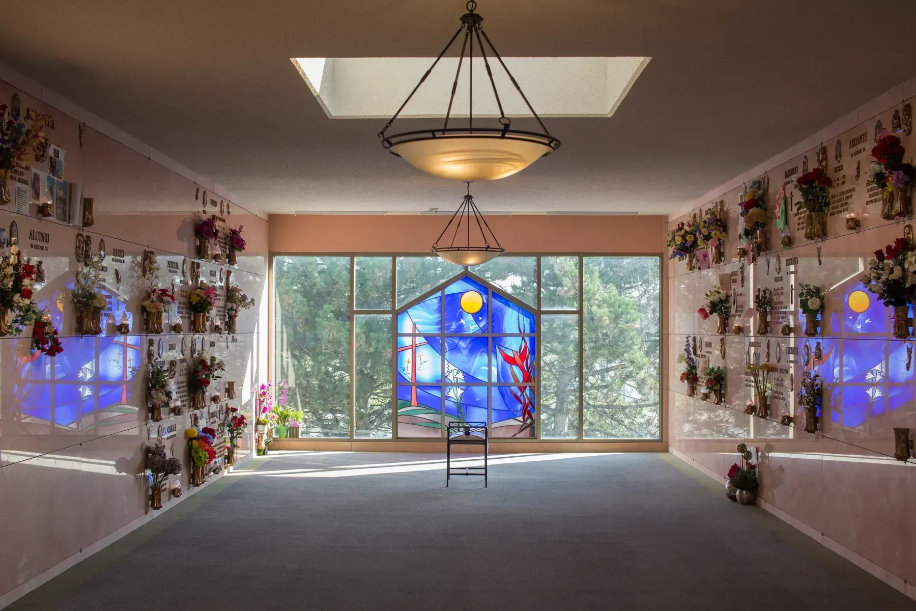 Flower-filled memorial room with blue stained-glass panels and a ladder centered by a large window