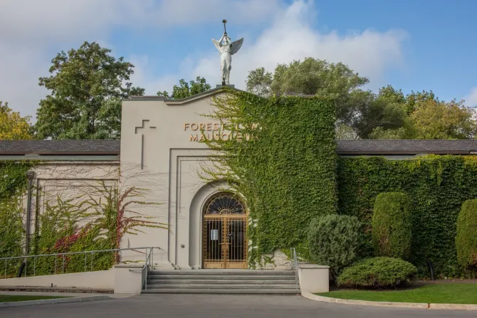 Stone building with arched entrance, cross, and ivy-covered walls surrounded by trees and greenery