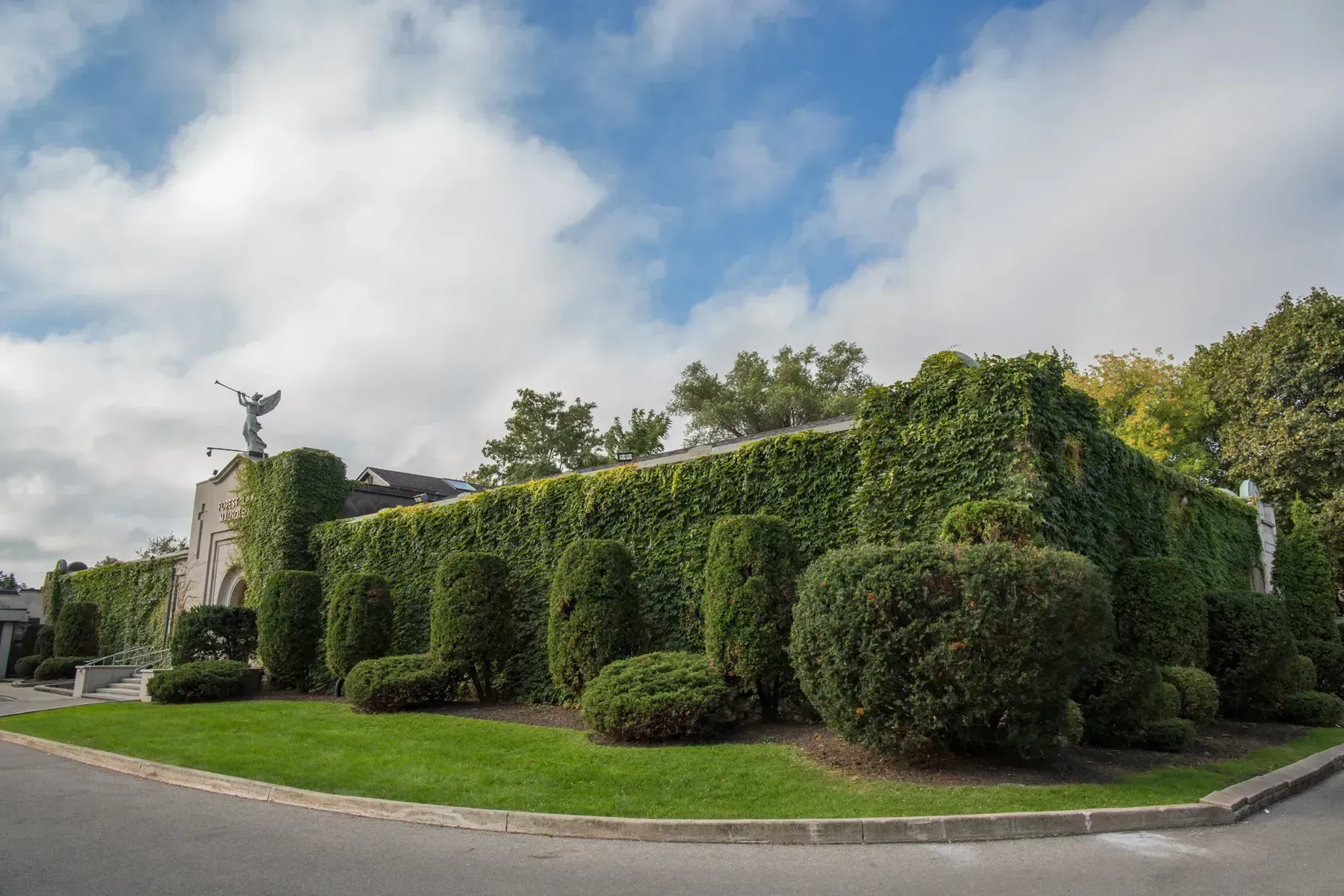 Dense green hedges form a building corner on a landscaped street under a partly cloudy sky
